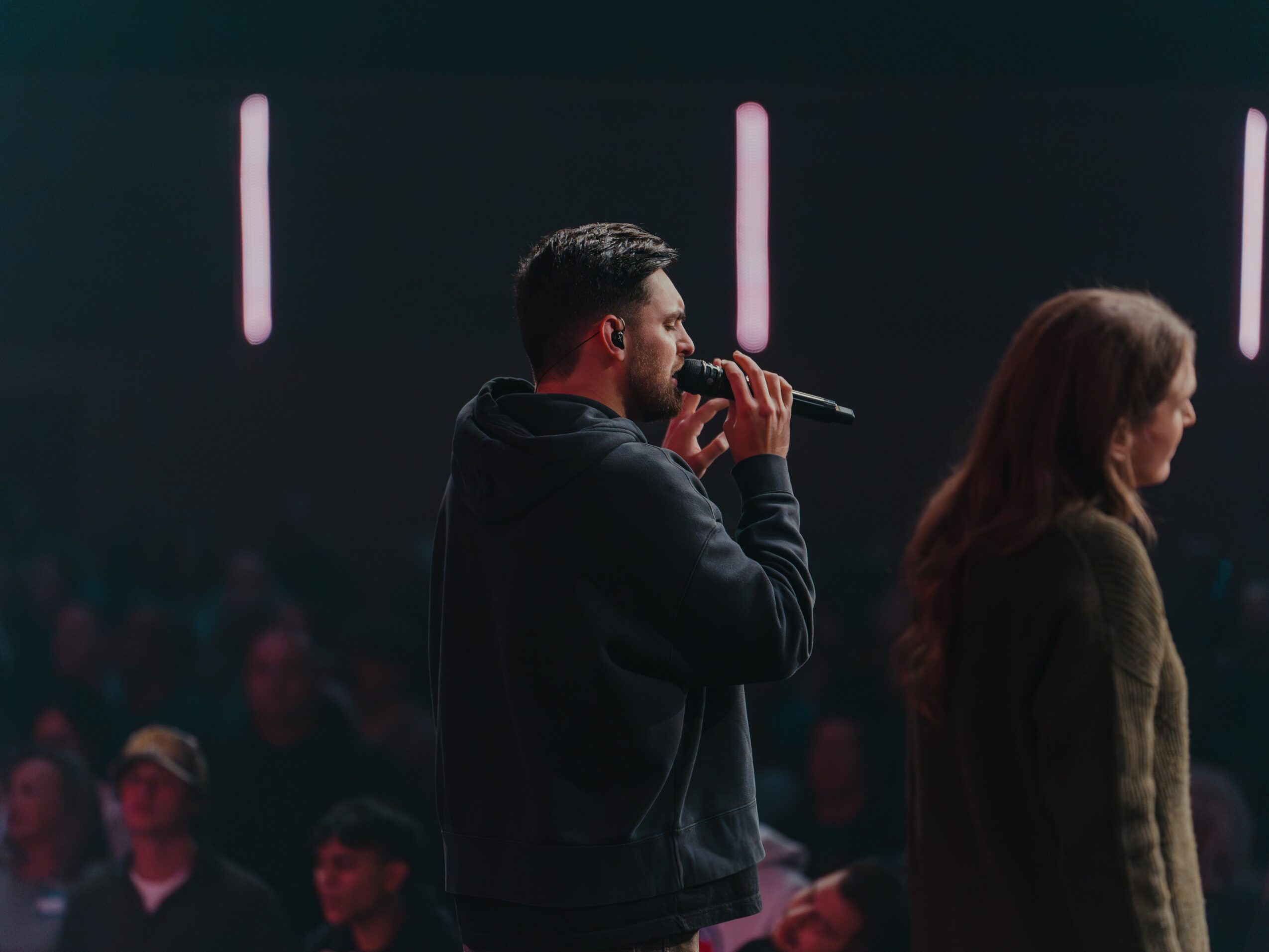 Man holding microphone speaking on stage with woman beside him, audience in background, dim lighting