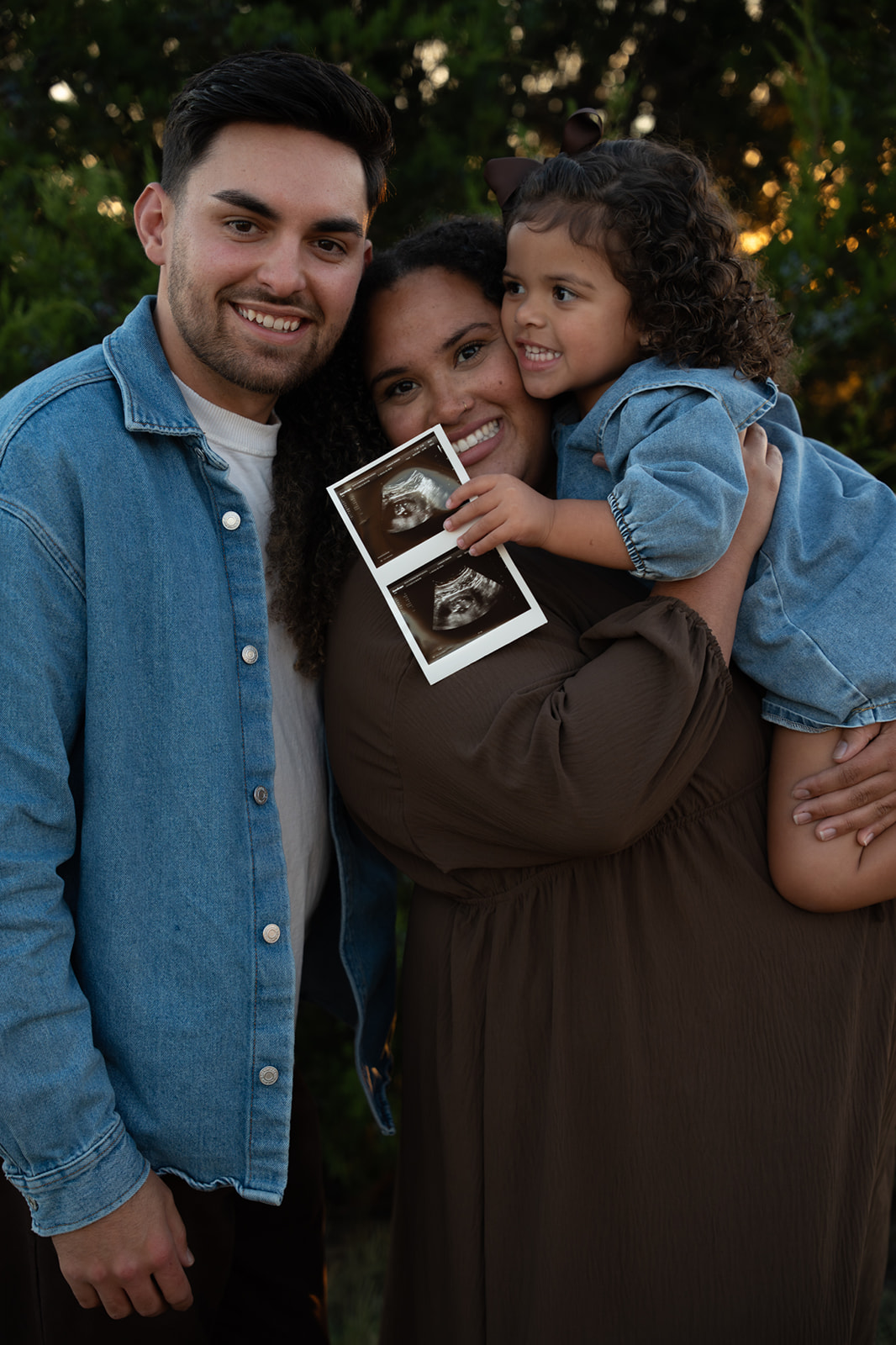 Family of three smiling outdoors, holding ultrasound images, with trees and lights in the background.