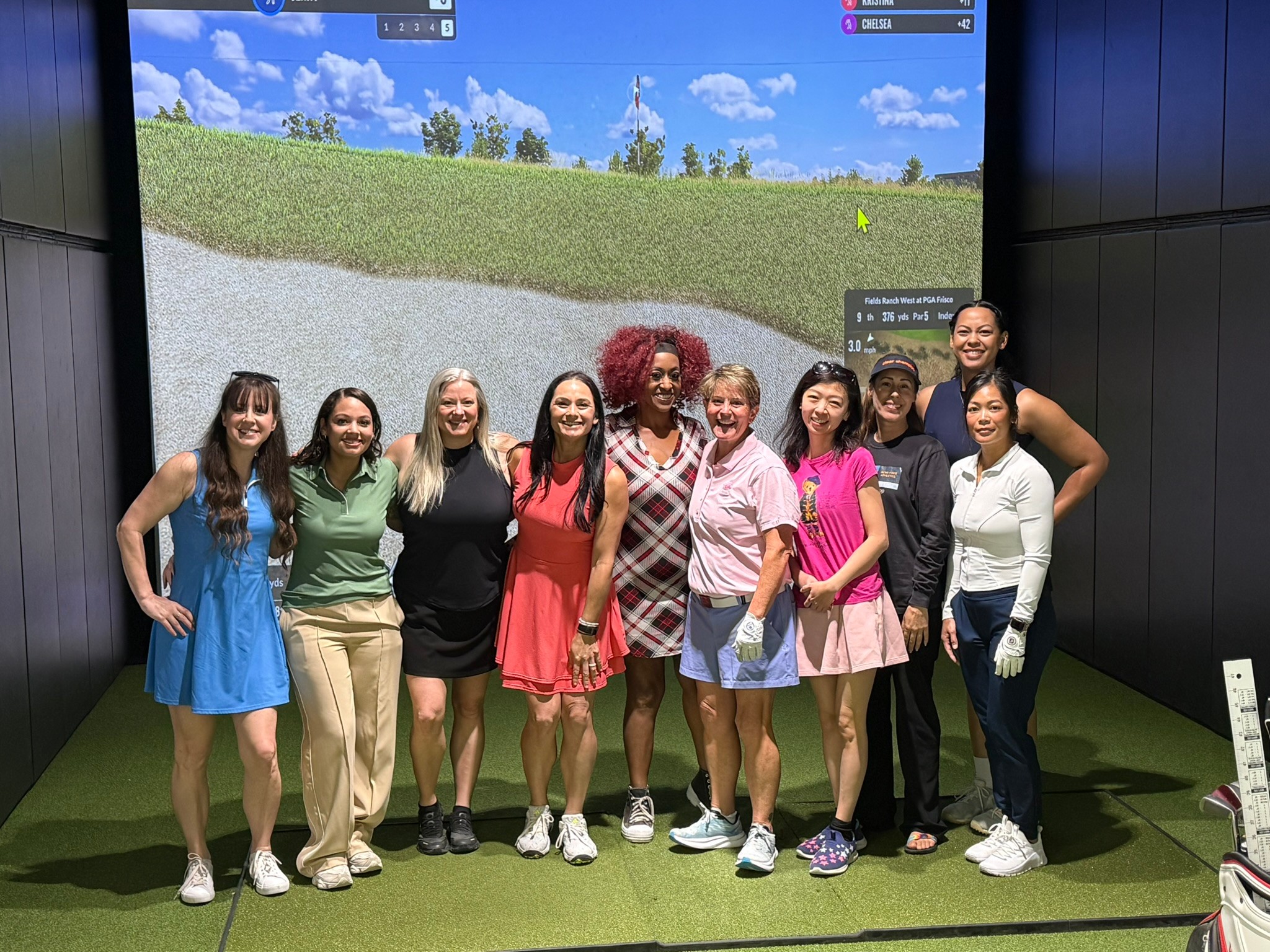Group of nine women standing indoors in front of a large screen displaying a golf course, smiling and posing for a photo.