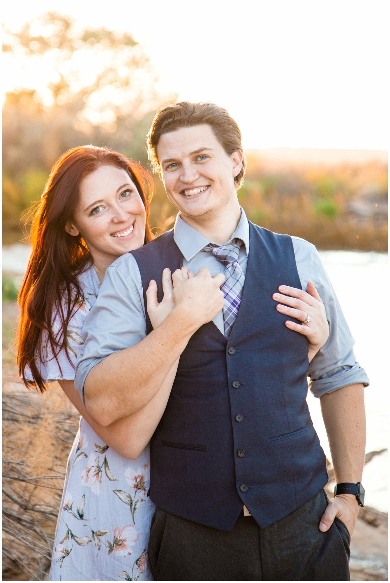 A smiling couple outdoors, the woman with red hair and the man in a vest, standing close together with sunlight background.