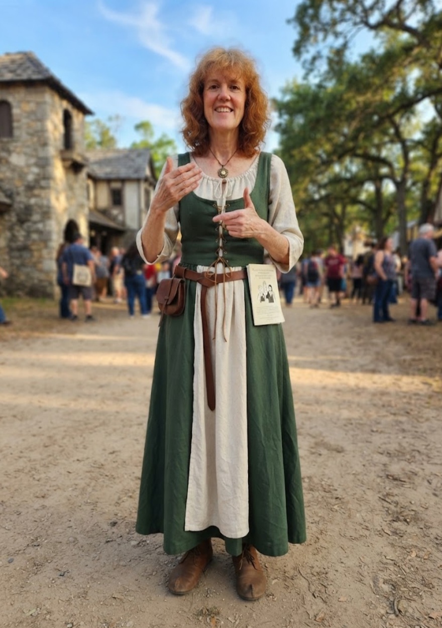 Woman with curly hair in medieval costume holding a cup outdoors, smiling, with people and trees in background.