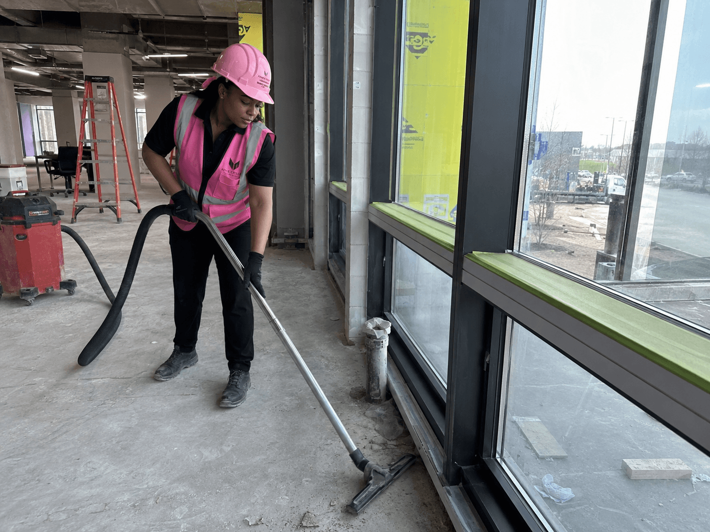Worker in pink safety helmet and vest vacuuming construction debris inside building near large windows.