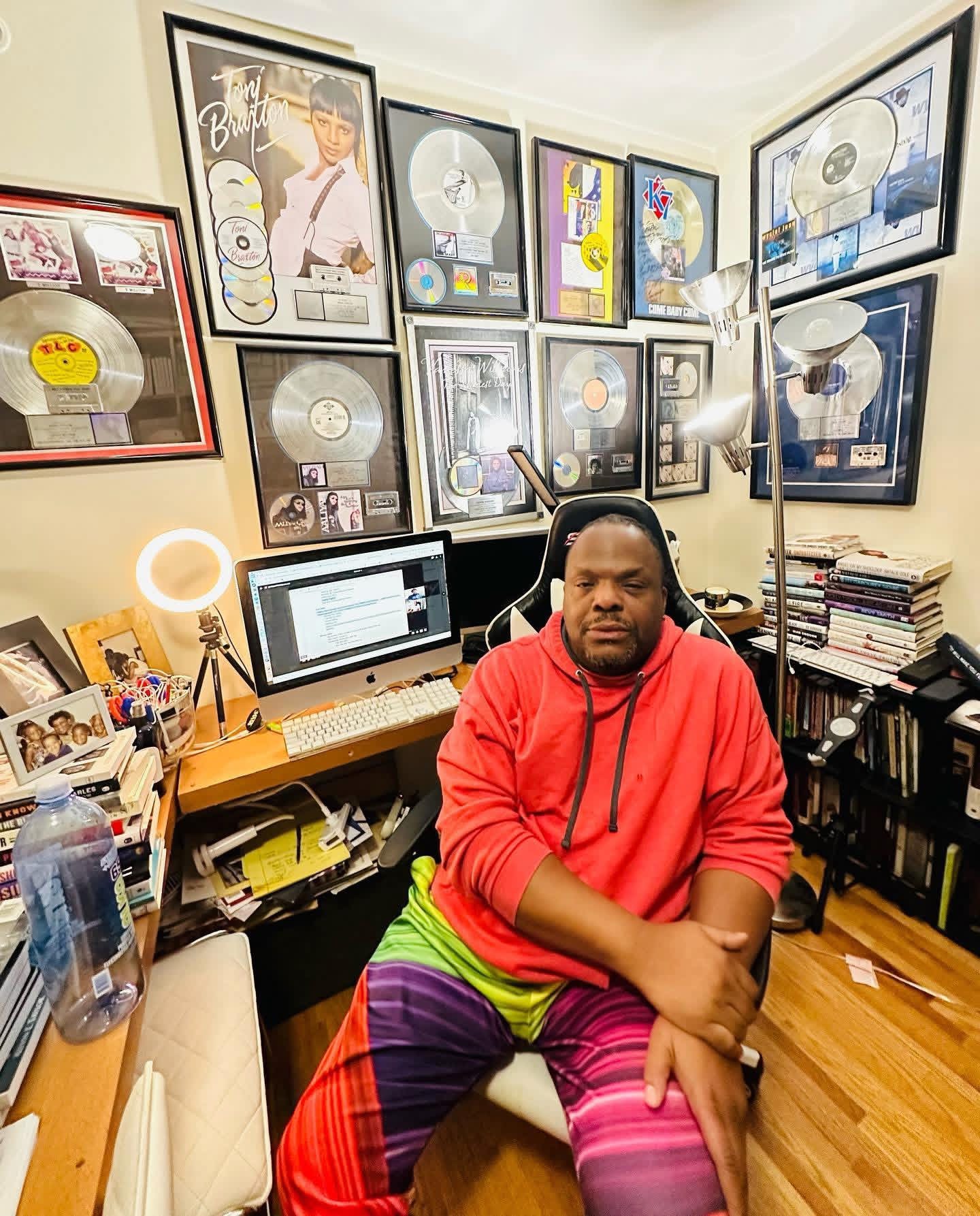 Man in red hoodie and colorful pants sitting in a room with framed music awards on the wall.