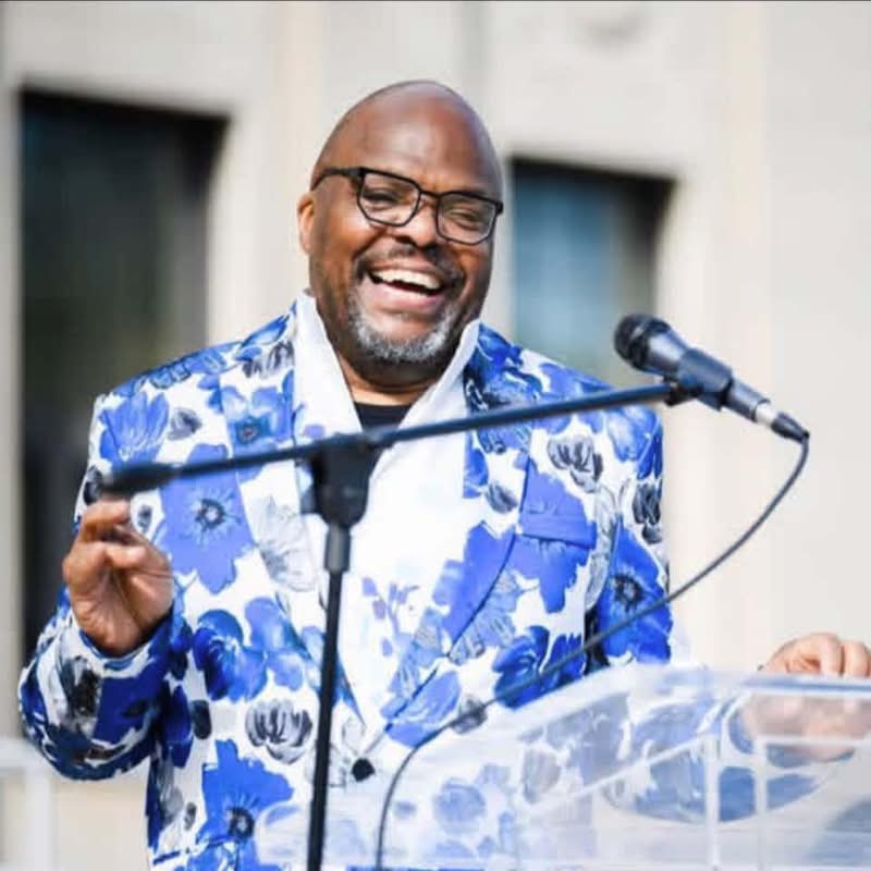 Man smiling, wearing glasses and a blue floral suit, speaking at a podium with a microphone outdoors.