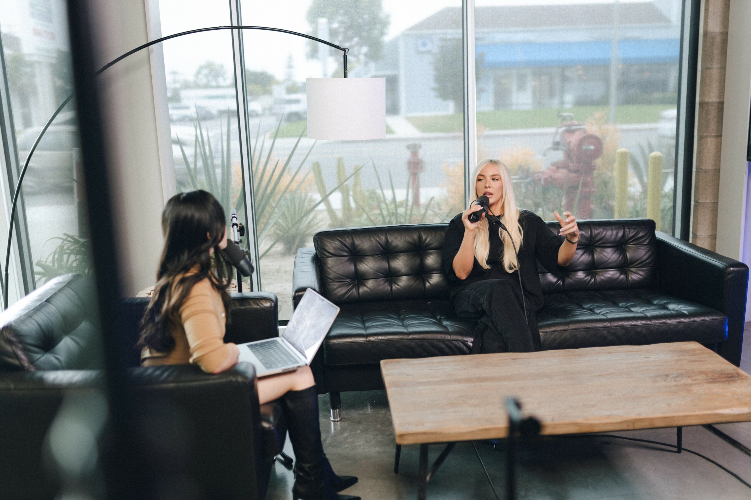 Two women sitting in a modern lounge area, one with a laptop and the other speaking into a microphone, with large windows behind them.
