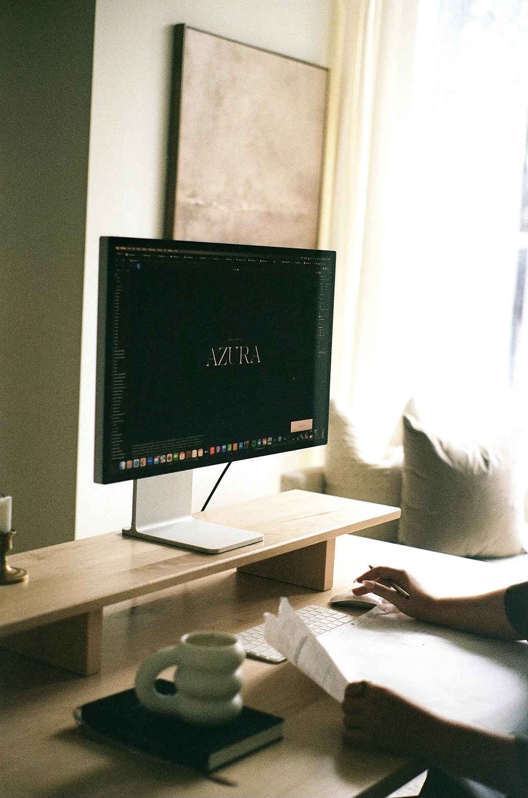 Person sitting on a sofa with a book, a computer monitor on a wooden shelf, and a window with curtains nearby.