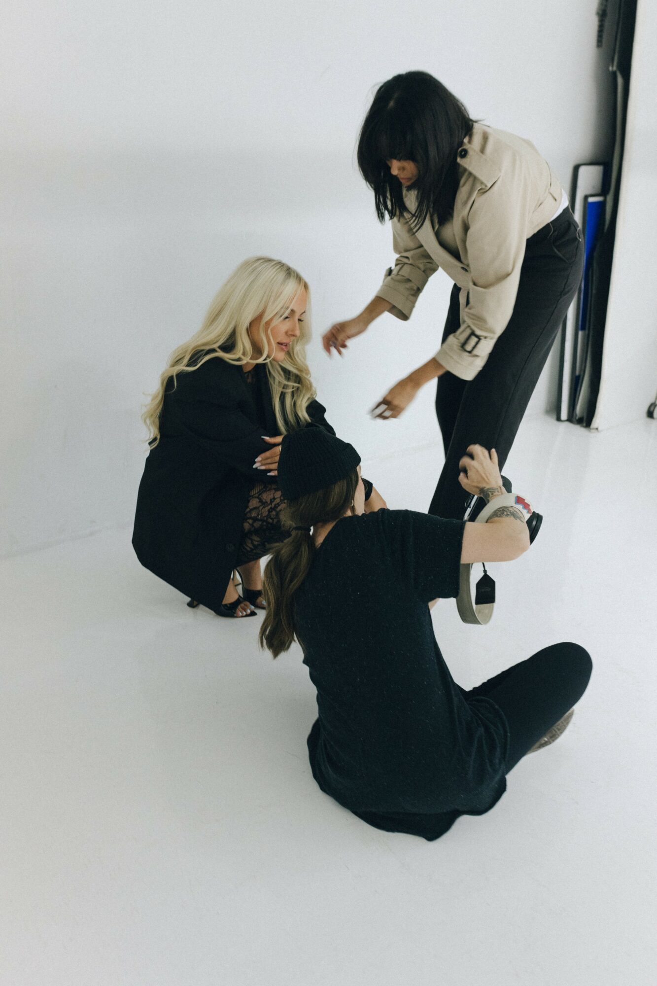 Three women are interacting on the floor in a white room, one sitting, one lying, and one standing, all engaged in conversation.