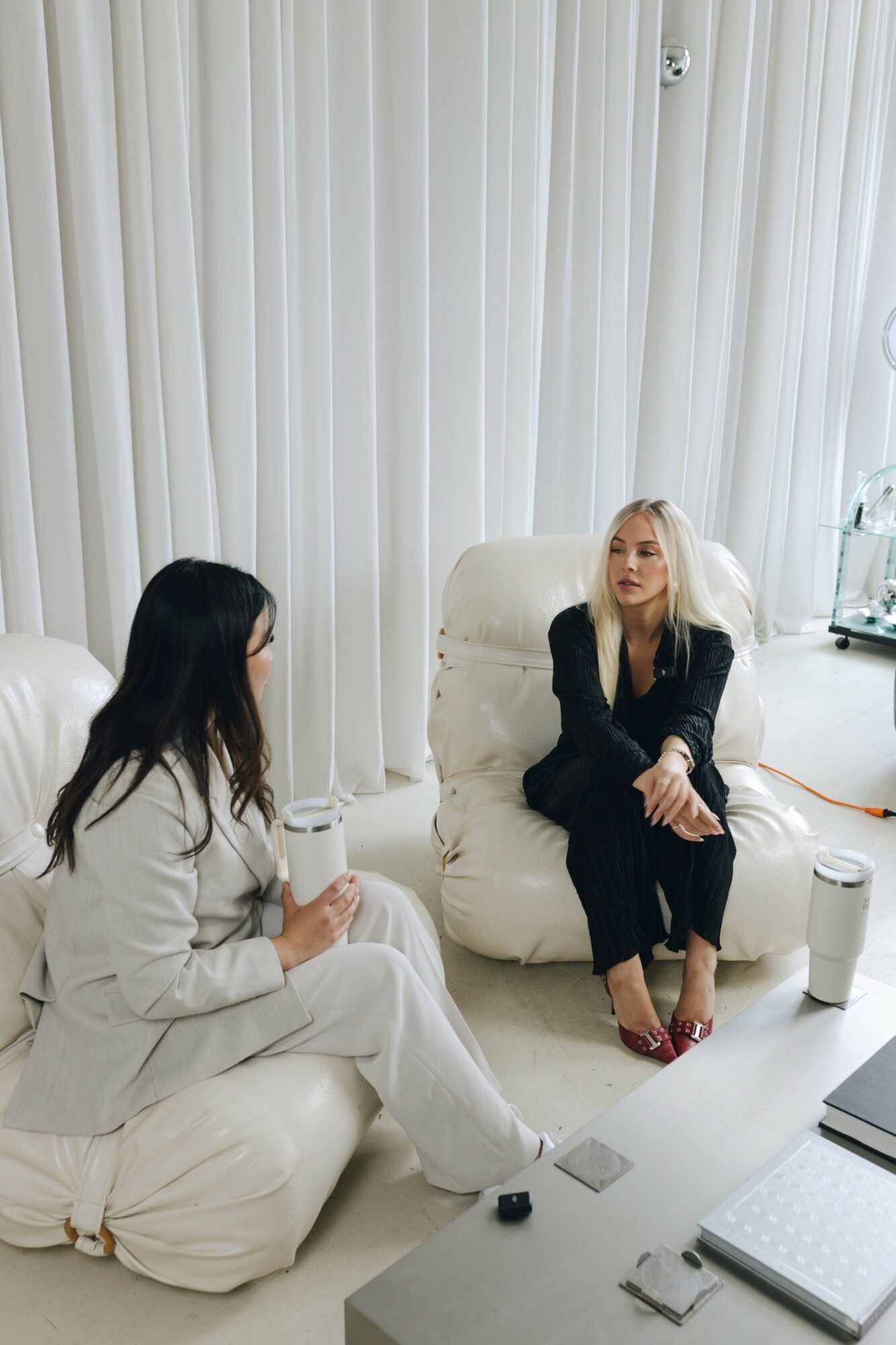 Two women sitting on white cushions in a room with white vertical blinds, one with long dark hair and the other with blonde hair, wearing black and white clothing.