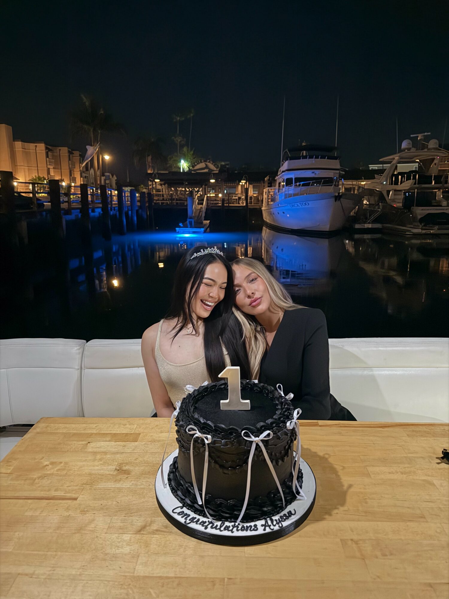 Two women sit together behind a birthday cake with a large number one candle, at a marina at night.