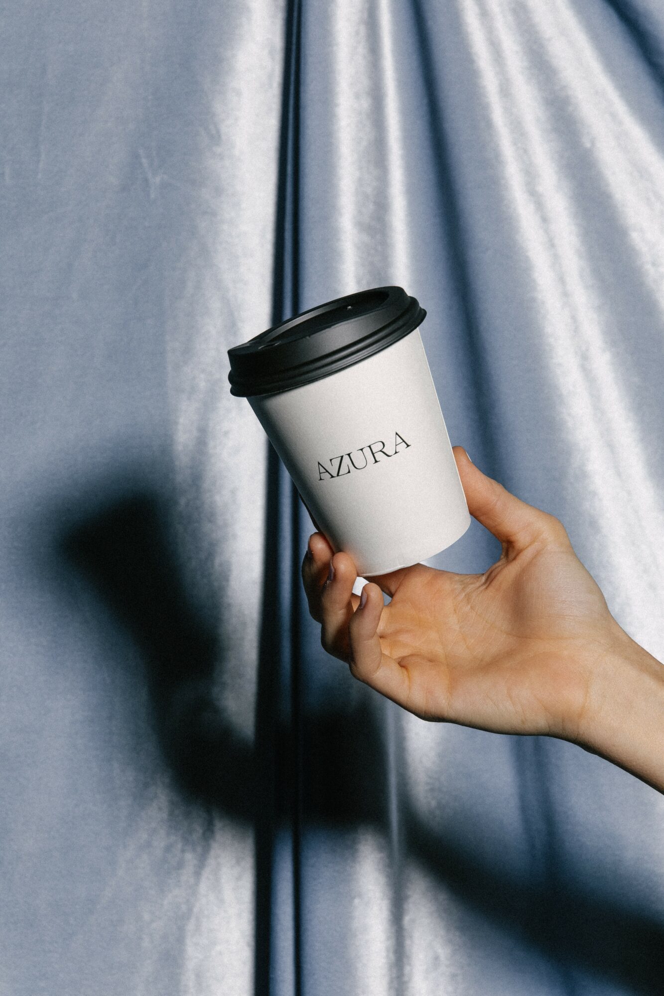 Hand holding a white coffee cup with black lid in front of shiny blue fabric background.