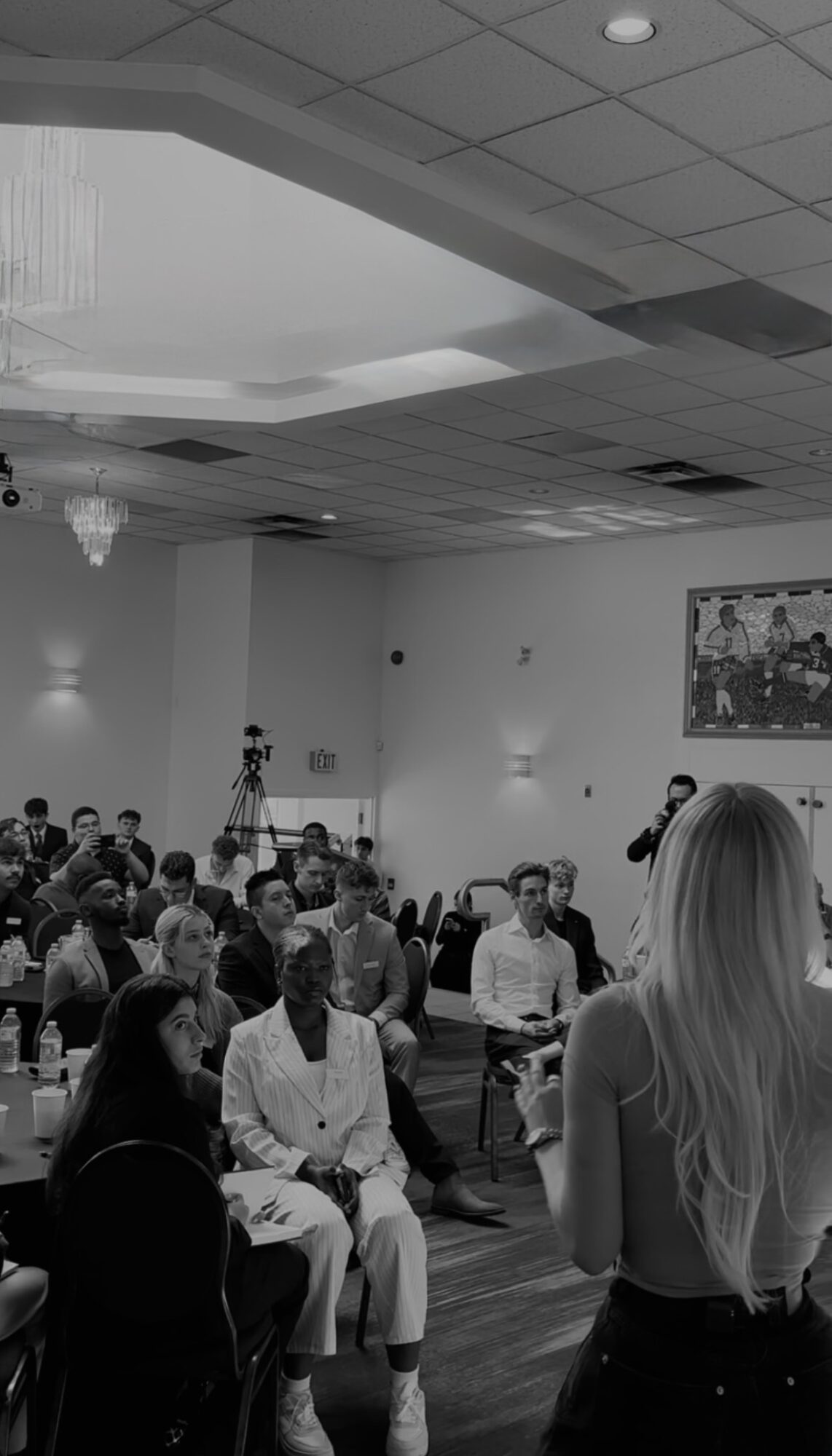 A woman with long hair speaking to a seated audience in a large room with high ceiling and artwork.
