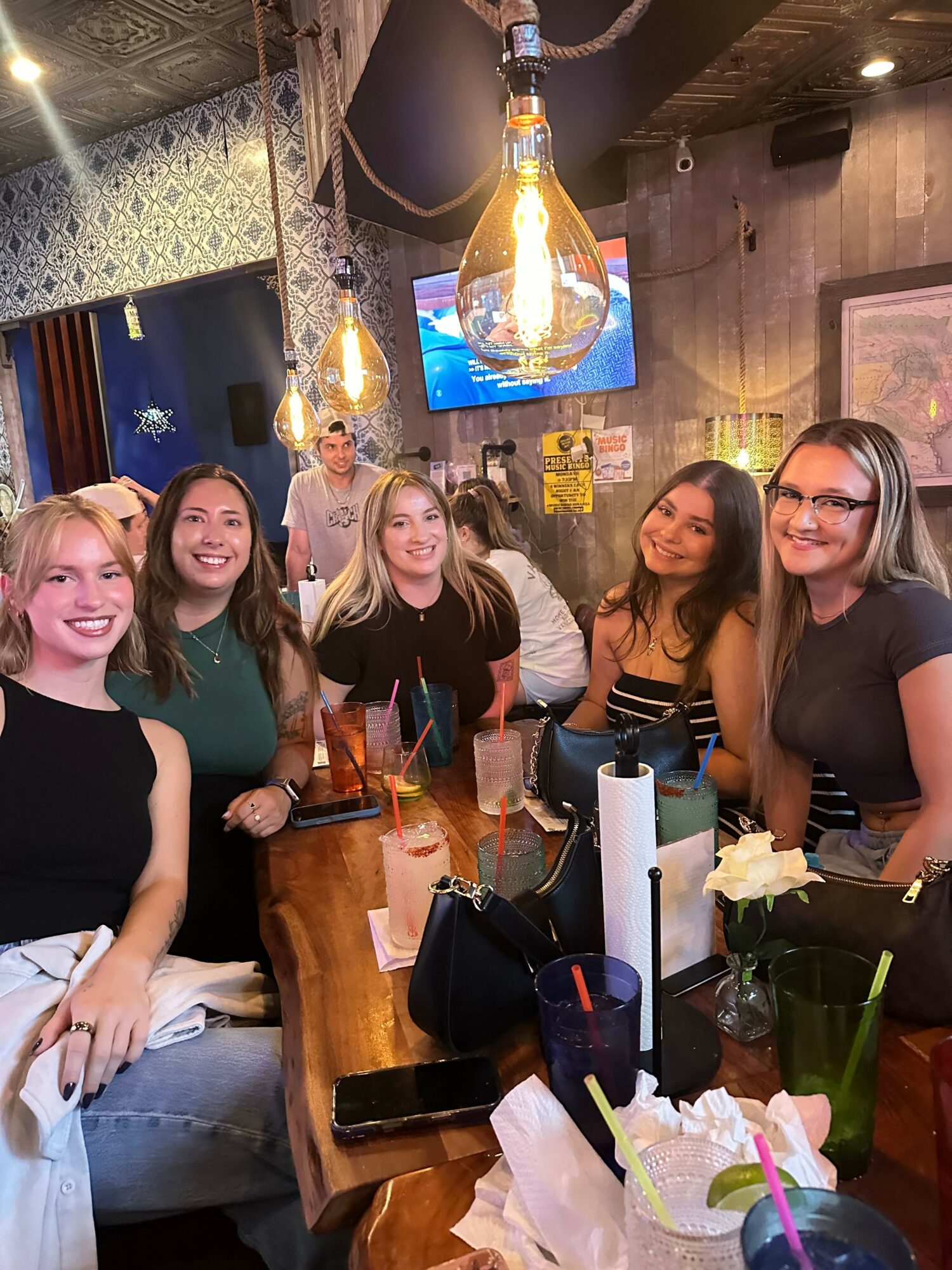 Group of six women sitting and standing around a table in a cozy, decorated indoor setting, smiling and enjoying drinks.