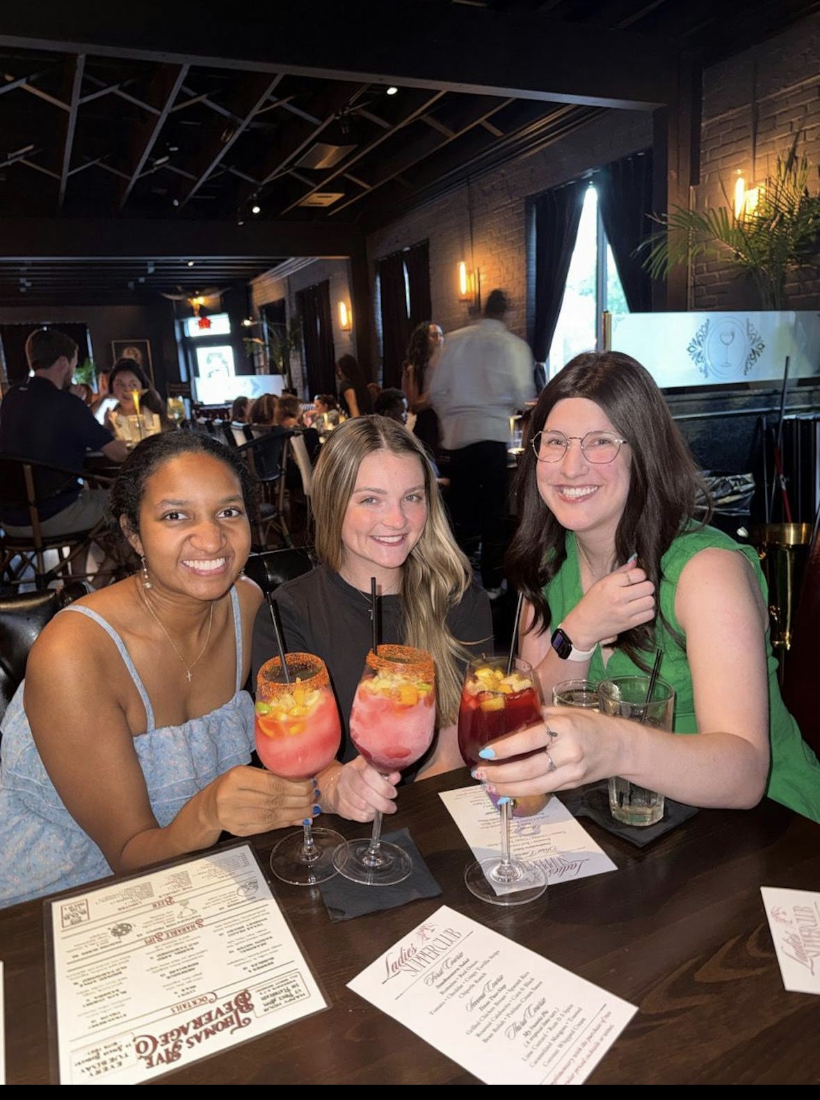 Three women sitting at a restaurant table holding colorful drinks, smiling, with menus and papers on the table.