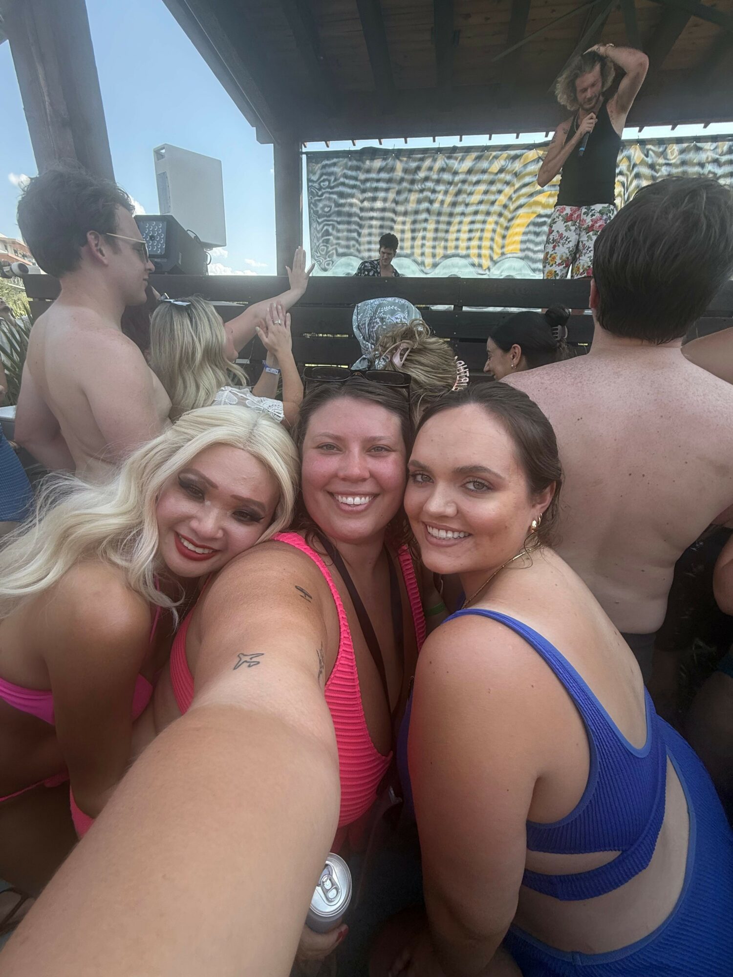 Three women smiling and taking a selfie together indoors, with a man in the background and a window showing a patterned wall.