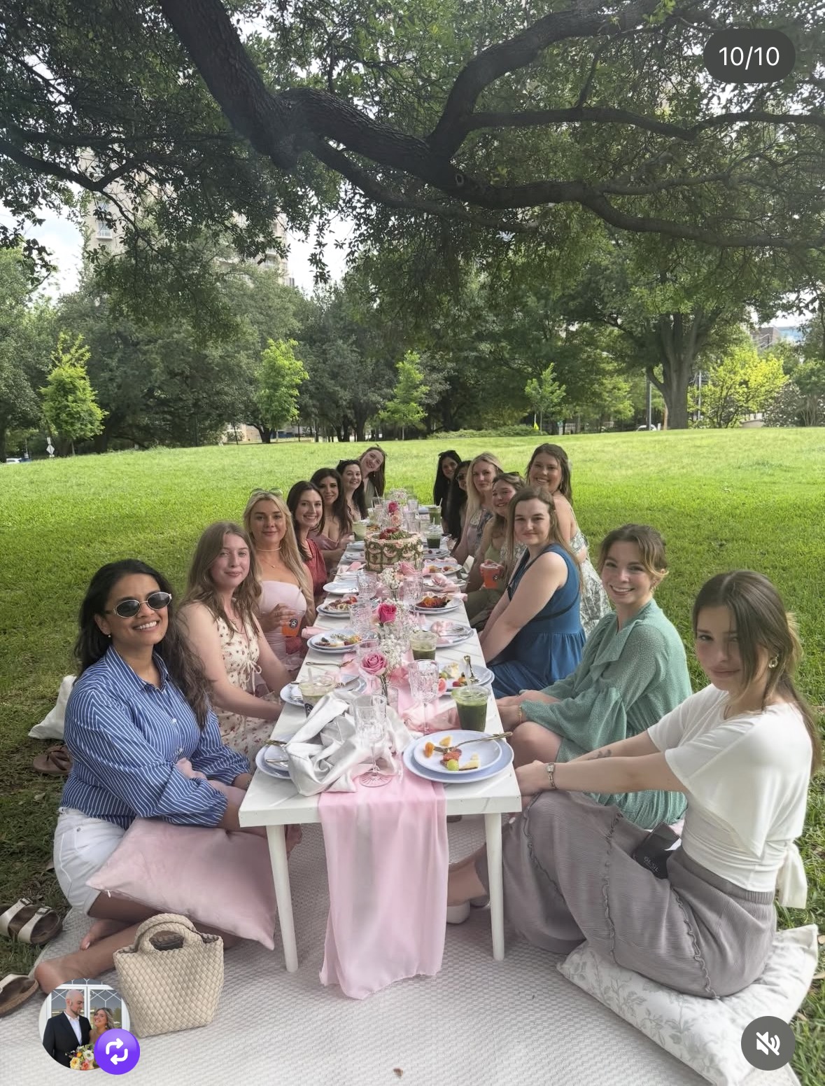 People sitting at a long table outdoors under a large tree, enjoying a meal with pink tablecloths and decorations.