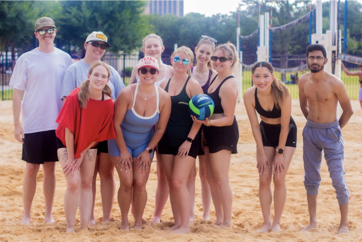 Group of people on a sandy volleyball court, some holding a volleyball, with trees and a building in the background.