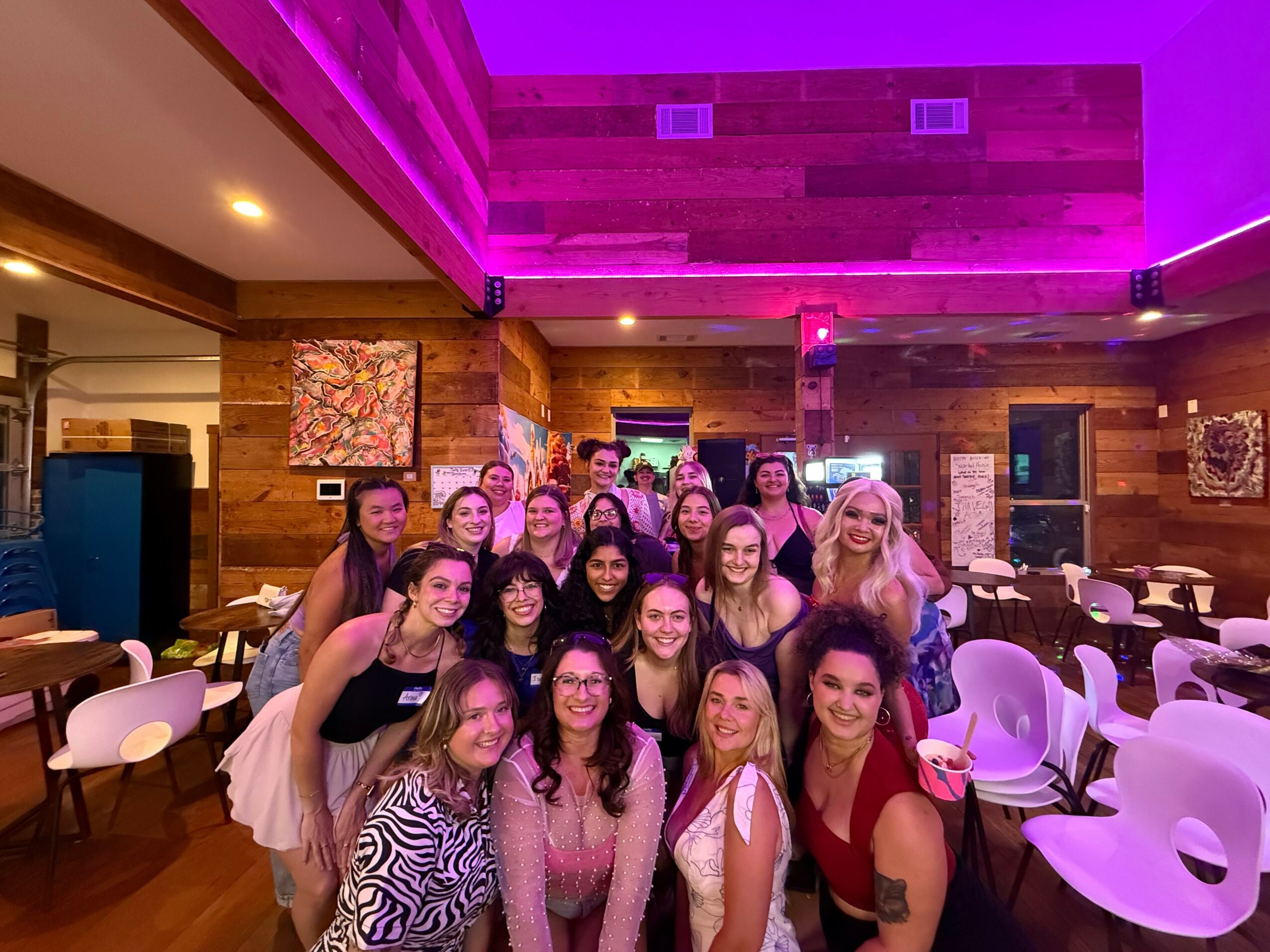 Group of women posing together in a decorated indoor venue with wooden walls and purple lighting, smiling and making gestures.
