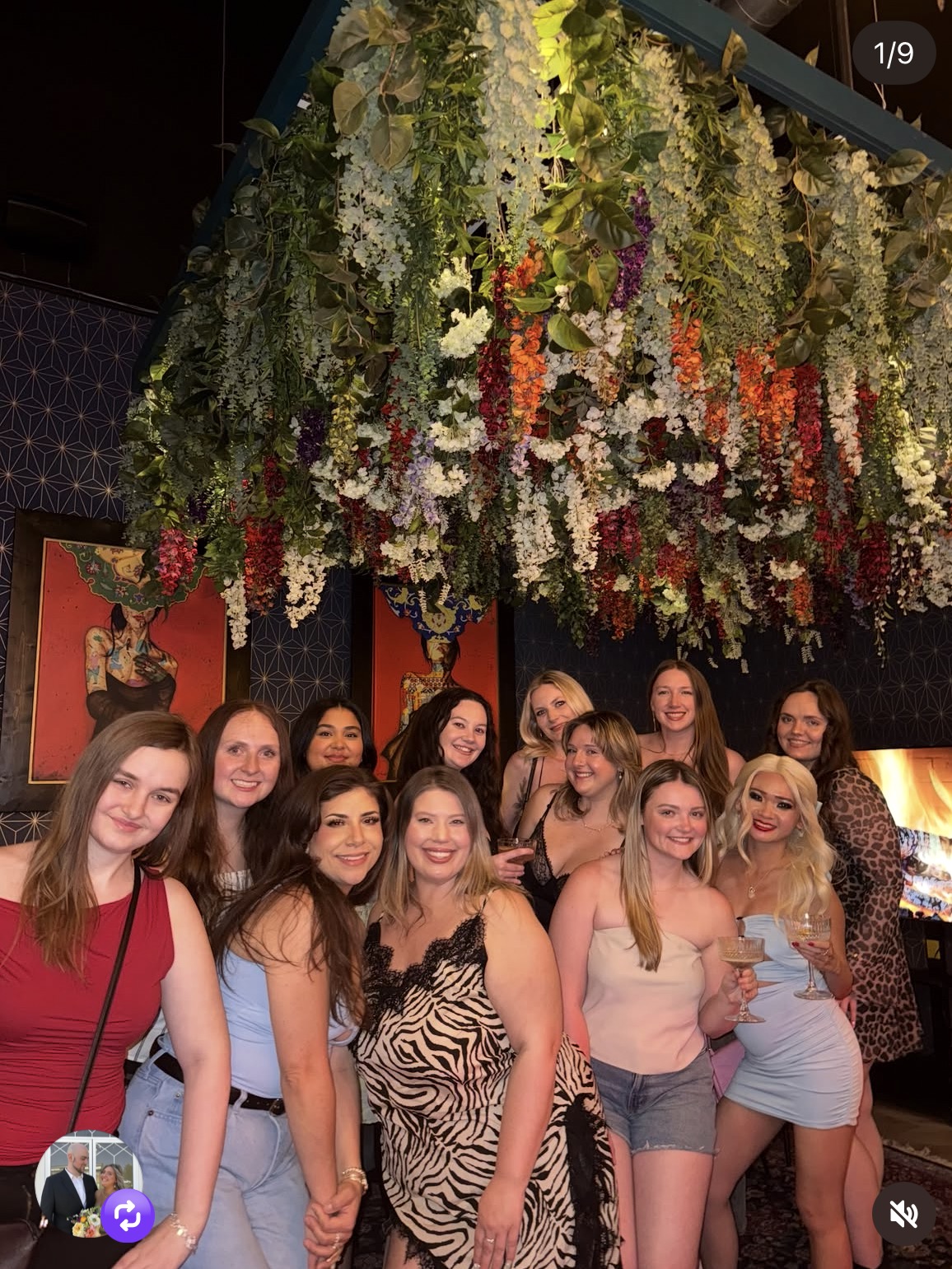 Group of women smiling indoors with hanging floral decorations above, dark walls, and framed artwork, celebrating together.