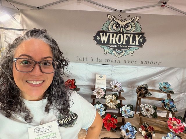 Woman with glasses smiling at a craft booth displaying colorful fabric flowers on wooden sticks.