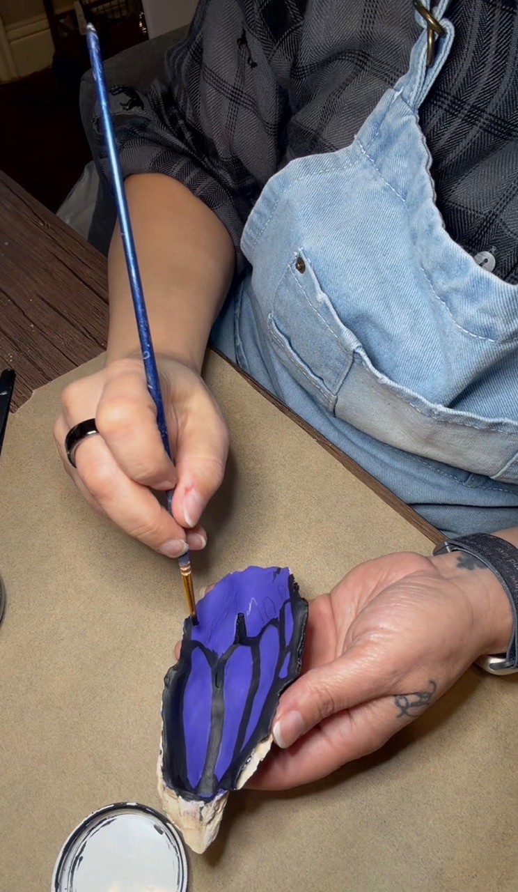 Person painting a butterfly-shaped object with blue and black colors, wearing a denim apron and black shirt.