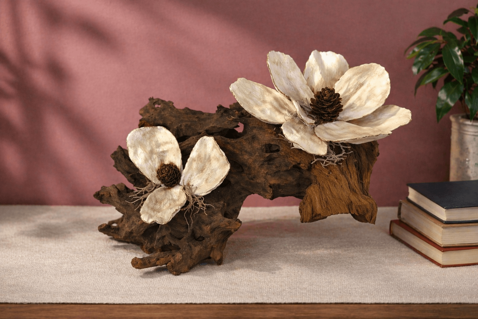 Decorative arrangement with large white flowers and dark centers on a piece of driftwood, with books and a plant in the background