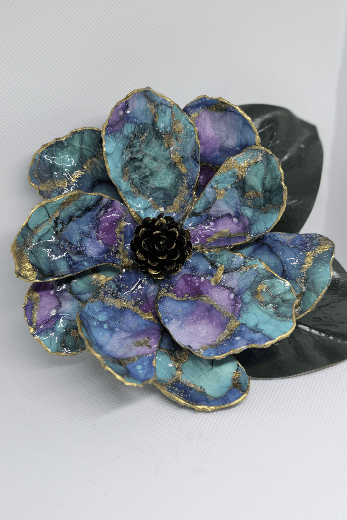Decorative flower with blue, purple, and gold accents, featuring a black center and dark green leaf, on a white background.