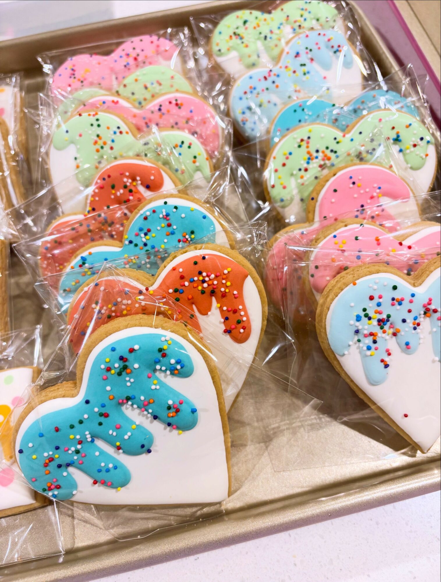 Heart-shaped cookies decorated with colorful icing and sprinkles, individually wrapped in plastic, arranged in a box.