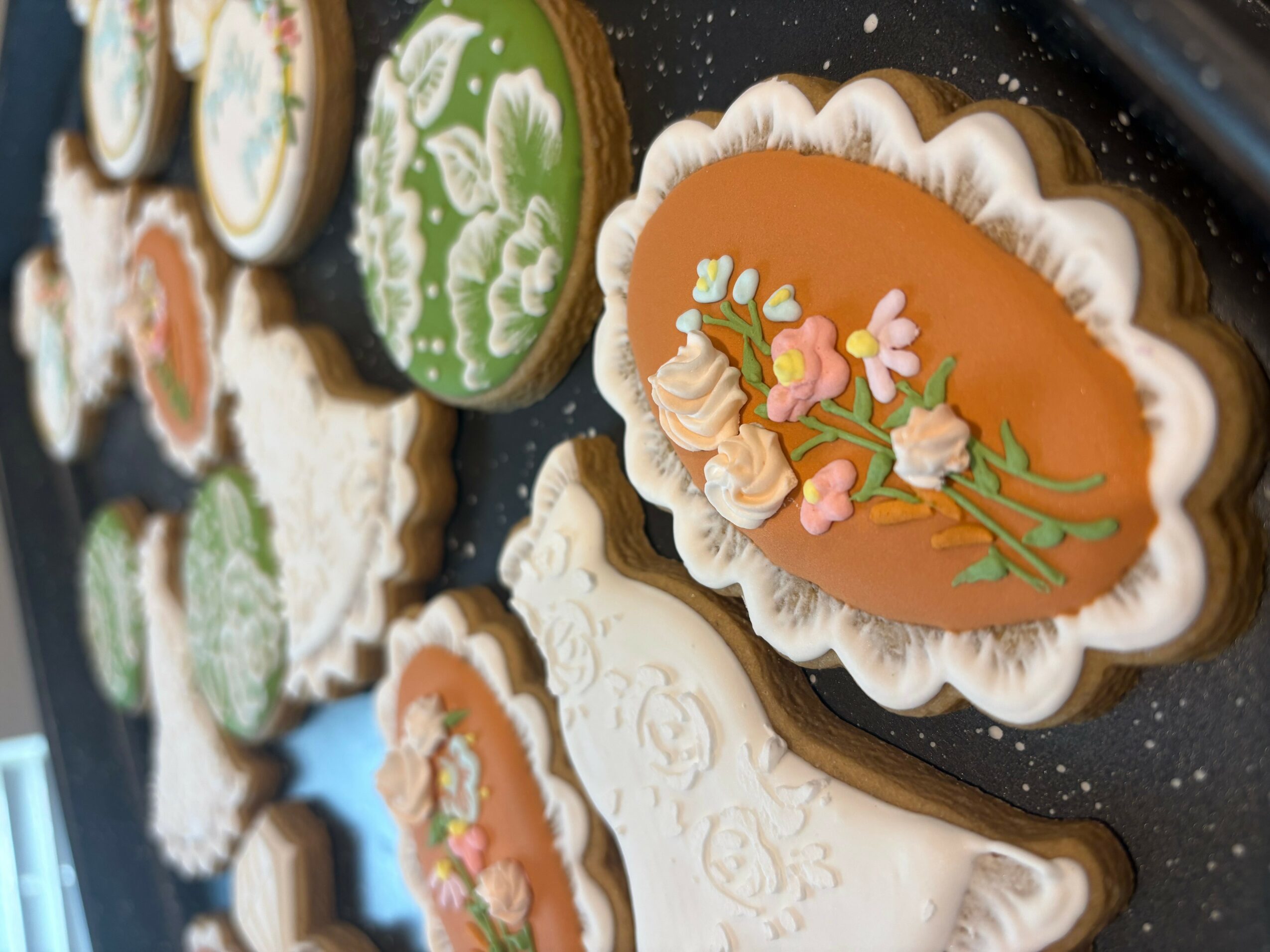 Decorated cookies with floral and ornate designs, some with icing flowers and intricate patterns, arranged on a black surface.