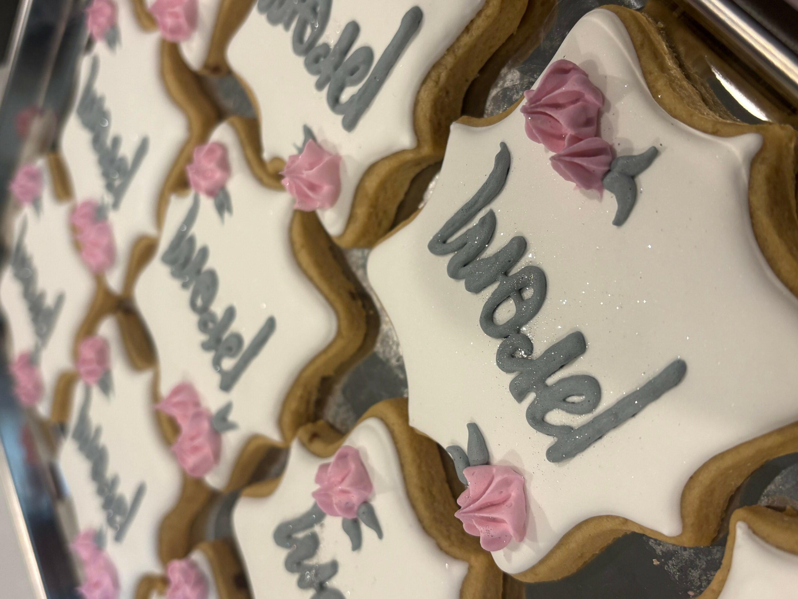 Decorative cookies with white icing, pink flowers, and gray writing, arranged in a pattern.