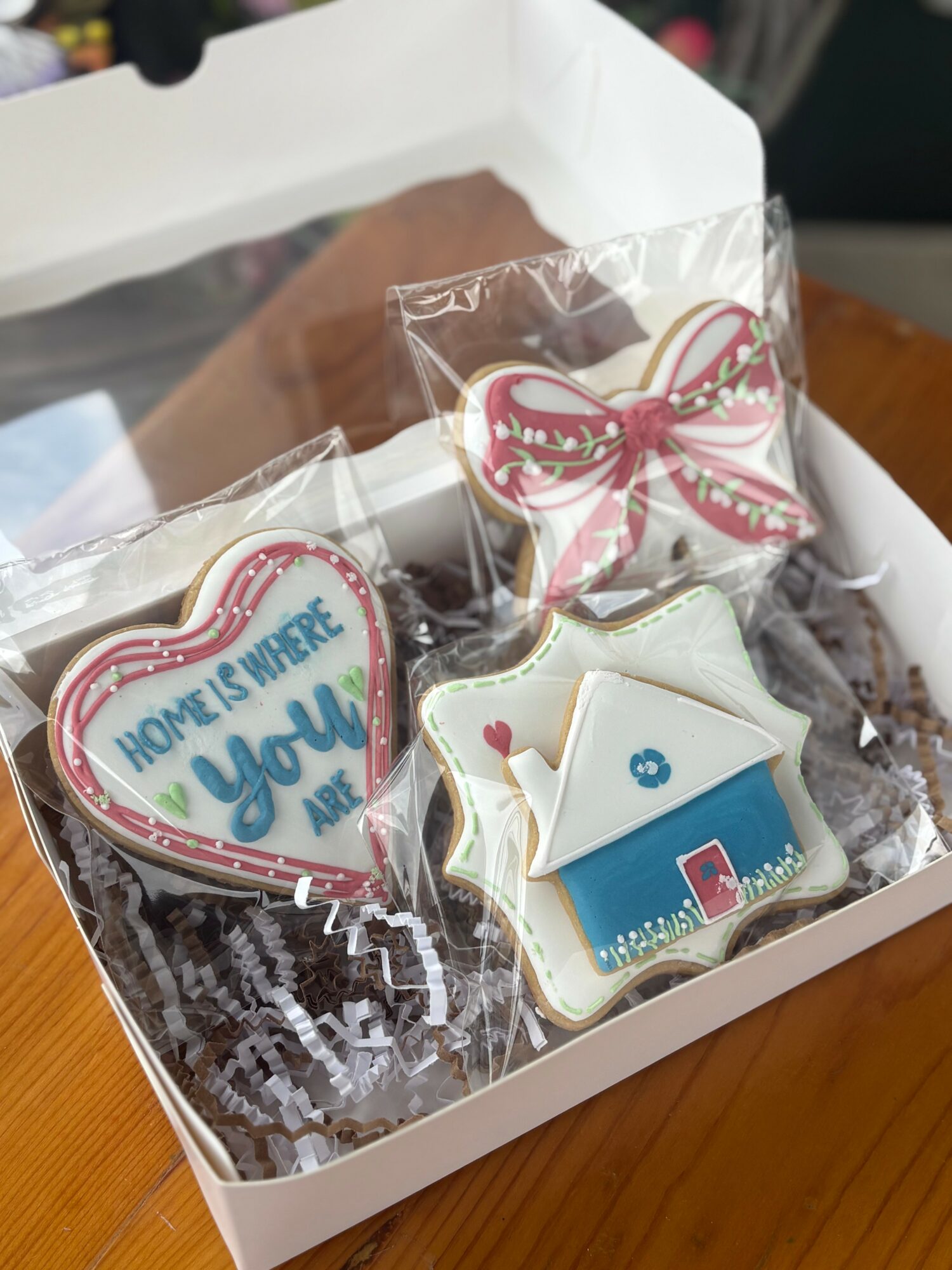 Decorated cookies in a box, including a heart with text, a bow, and a house, on shredded paper filling.