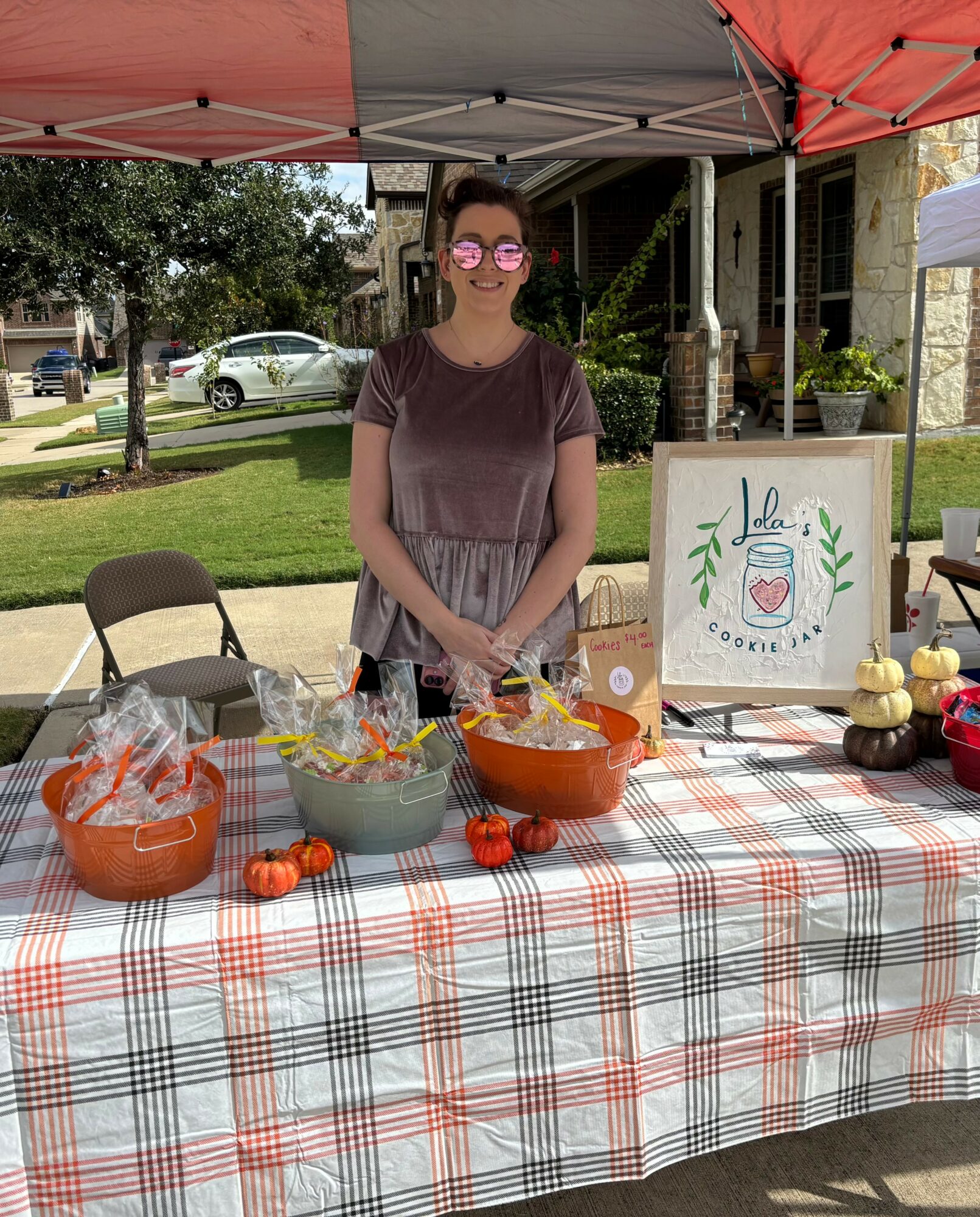 Young woman standing behind a table with baked goods and a sign, outdoors on a sunny day.