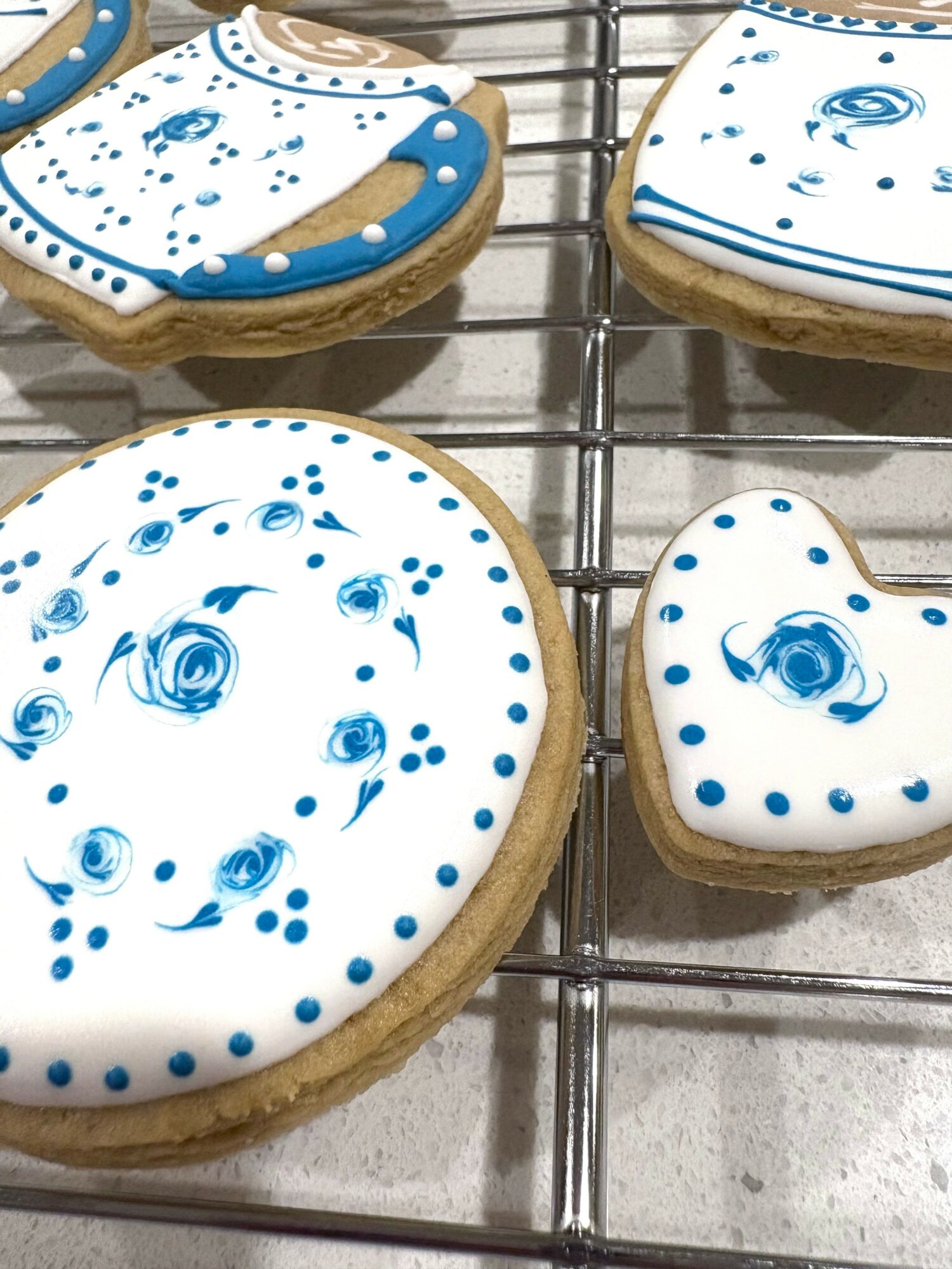 Four cookies decorated with blue and white icing, placed on a wire rack. Two cookies are round, two are heart-shaped.