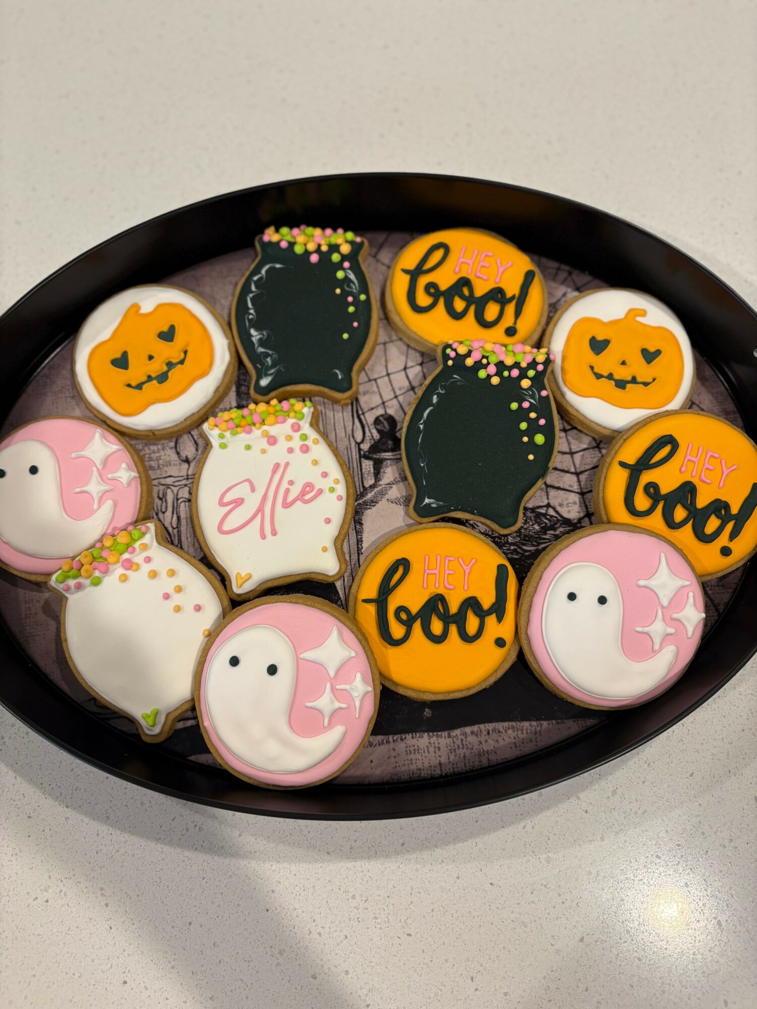 Halloween-themed decorated cookies on a black tray, featuring pumpkins, ghosts, and the word 'boo!', with colorful sprinkles.