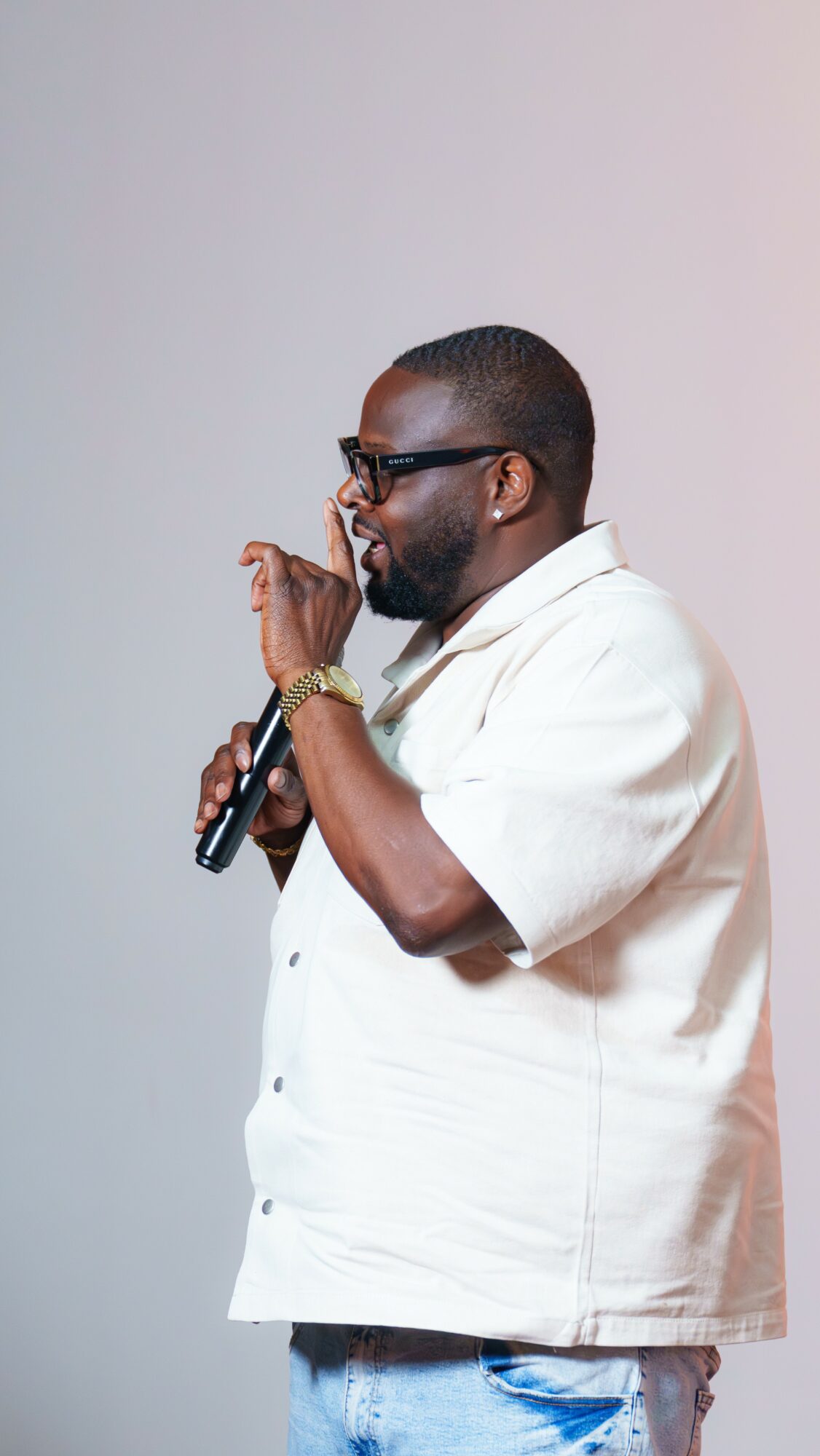 Man with glasses and beard holding a microphone, wearing a white shirt and jeans, standing sideways against a plain background.