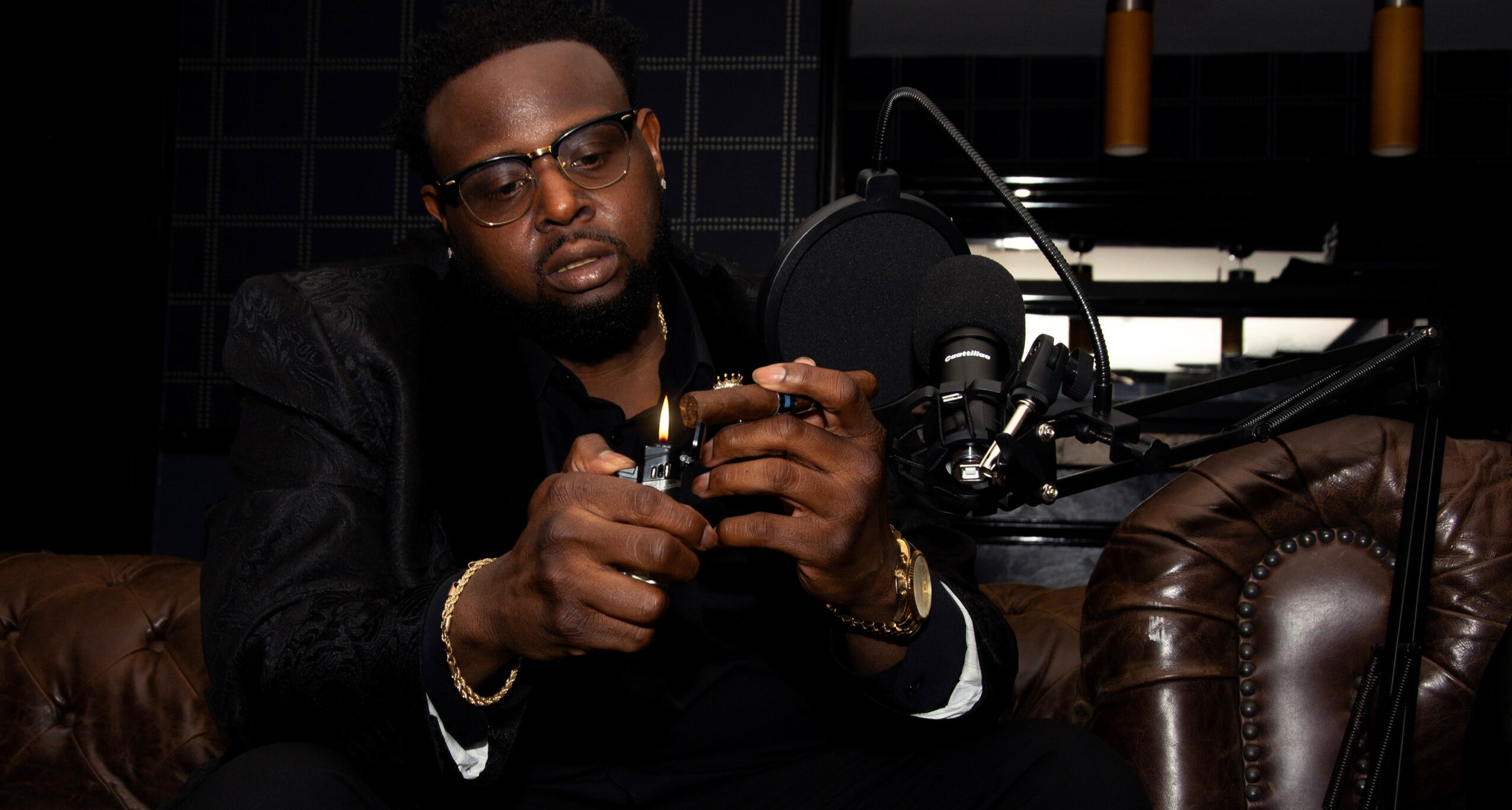 Man with glasses and a beard, sitting in a dark room, holding a small object, with a microphone nearby.