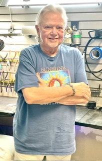 Elderly woman with short white hair smiling, arms crossed, in a workshop with tools and equipment behind her.