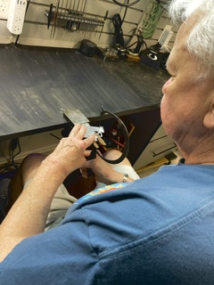 Elderly person working on a small electronic device at a cluttered workbench, with tools and supplies in the background.