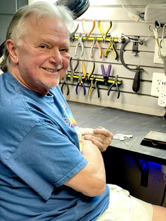 Older man with gray hair smiling, sitting in a workshop with tools hanging on the wall behind him.
