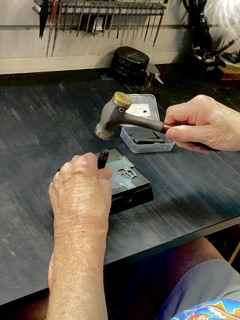 Person holding a tool over a black work surface with various tools and objects in the background.