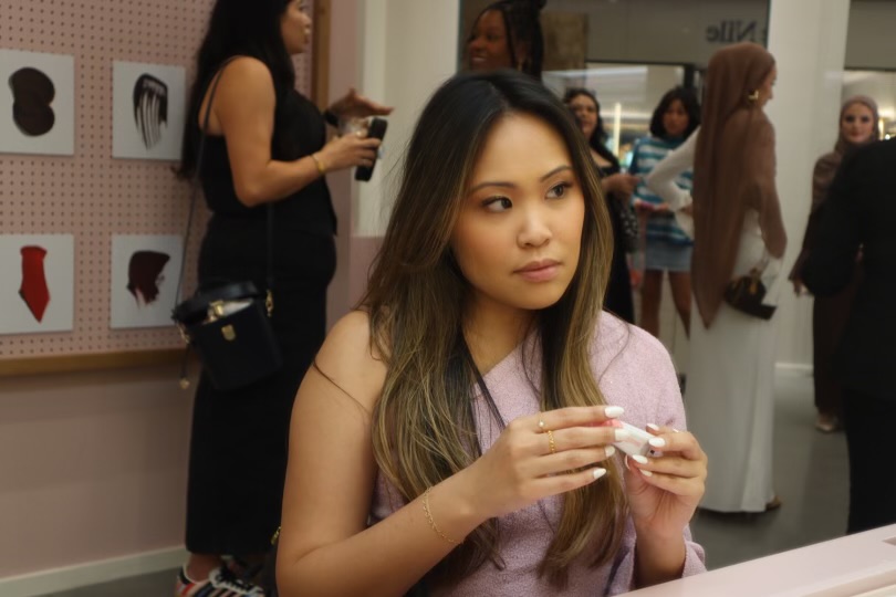 Young woman with long hair holding a small object, in a room with people and display boards, casual setting.