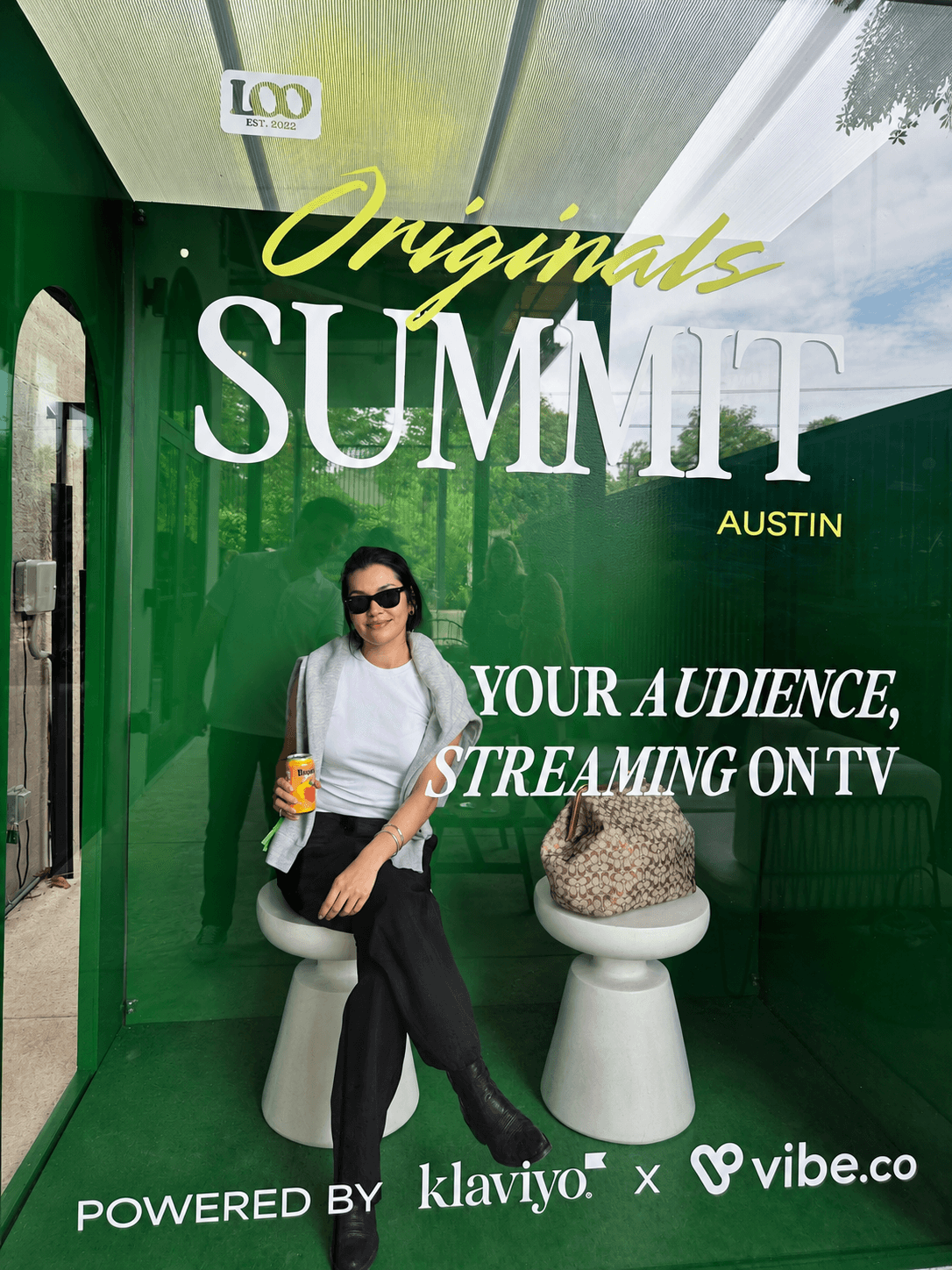 Woman sitting on a stool holding a drink, green background with event text, sunglasses, casual outfit, bag nearby.