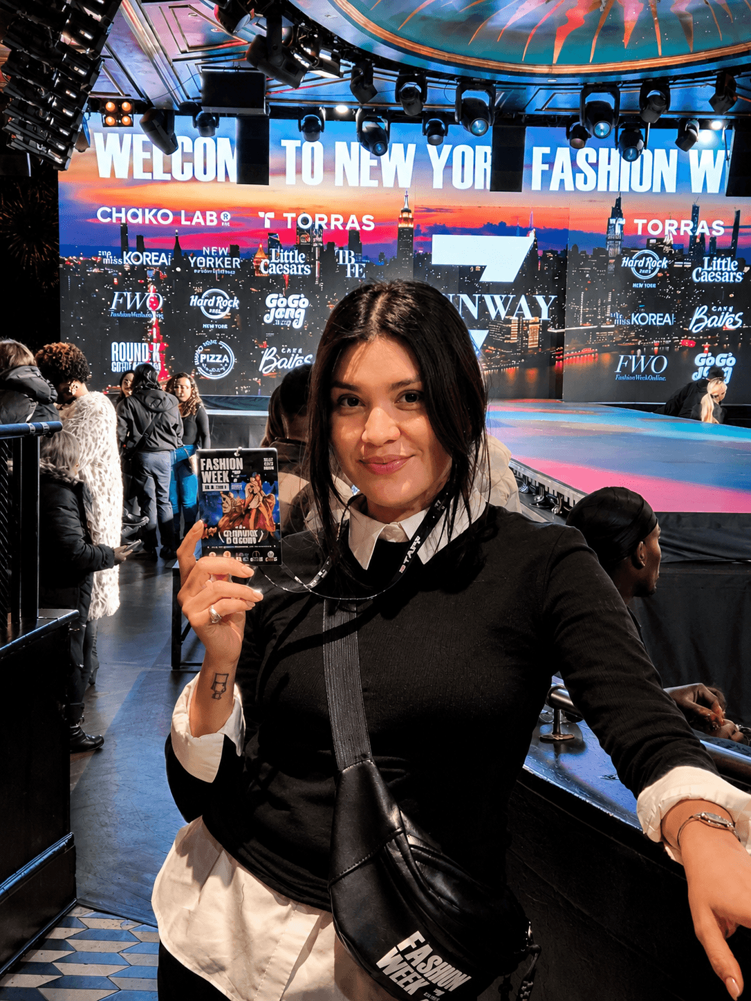 Woman holding a small object on stage at a fashion event with a large screen behind her displaying event information.