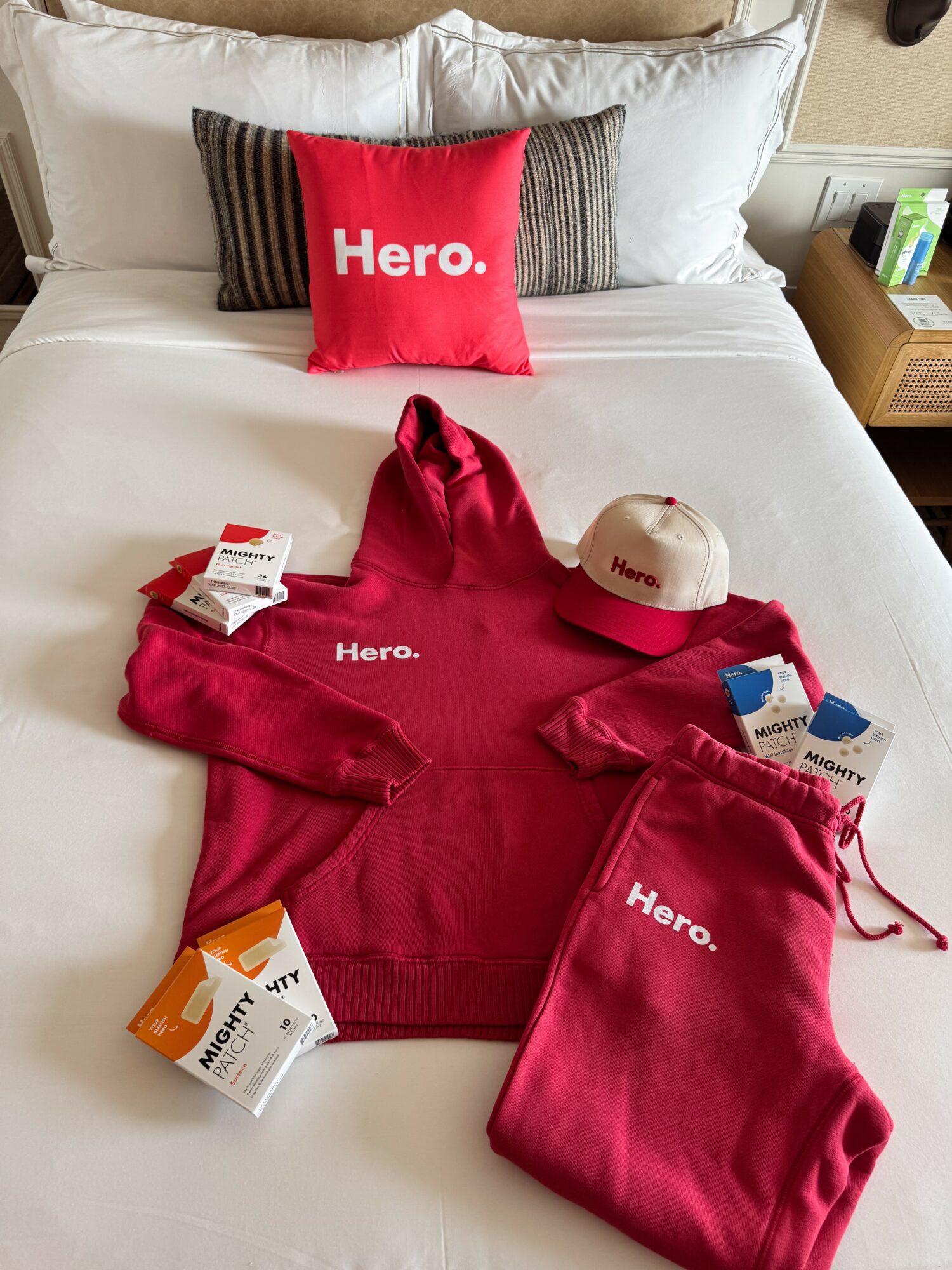 Red Hero hoodie, cap, and sweatpants on bed with pillows, tags, and a bedside table.