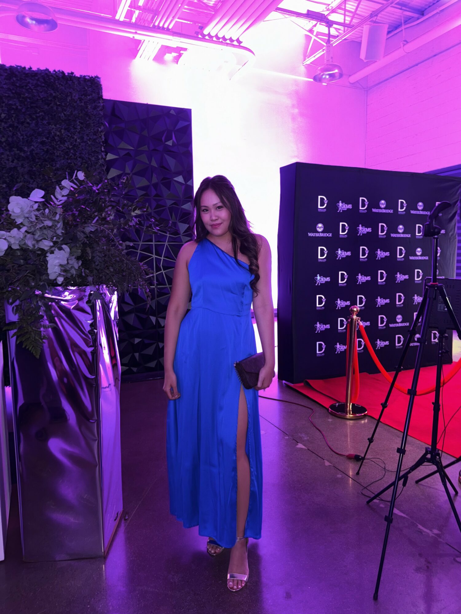 Woman in blue dress standing indoors with purple lighting, next to a black backdrop and red carpet, holding a clutch.
