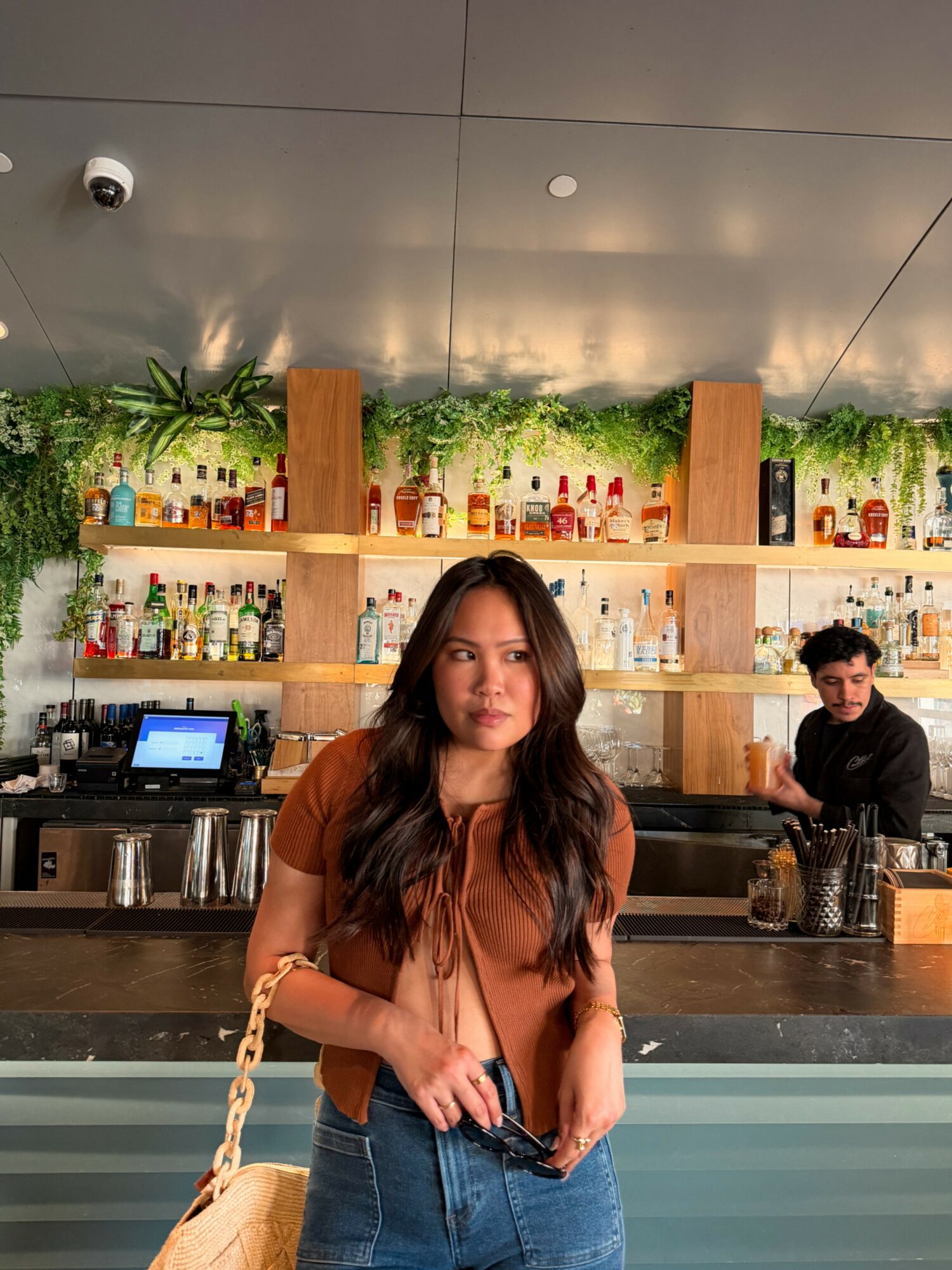 Woman with long hair standing at bar counter with shelves of bottles behind her, bartender in background.