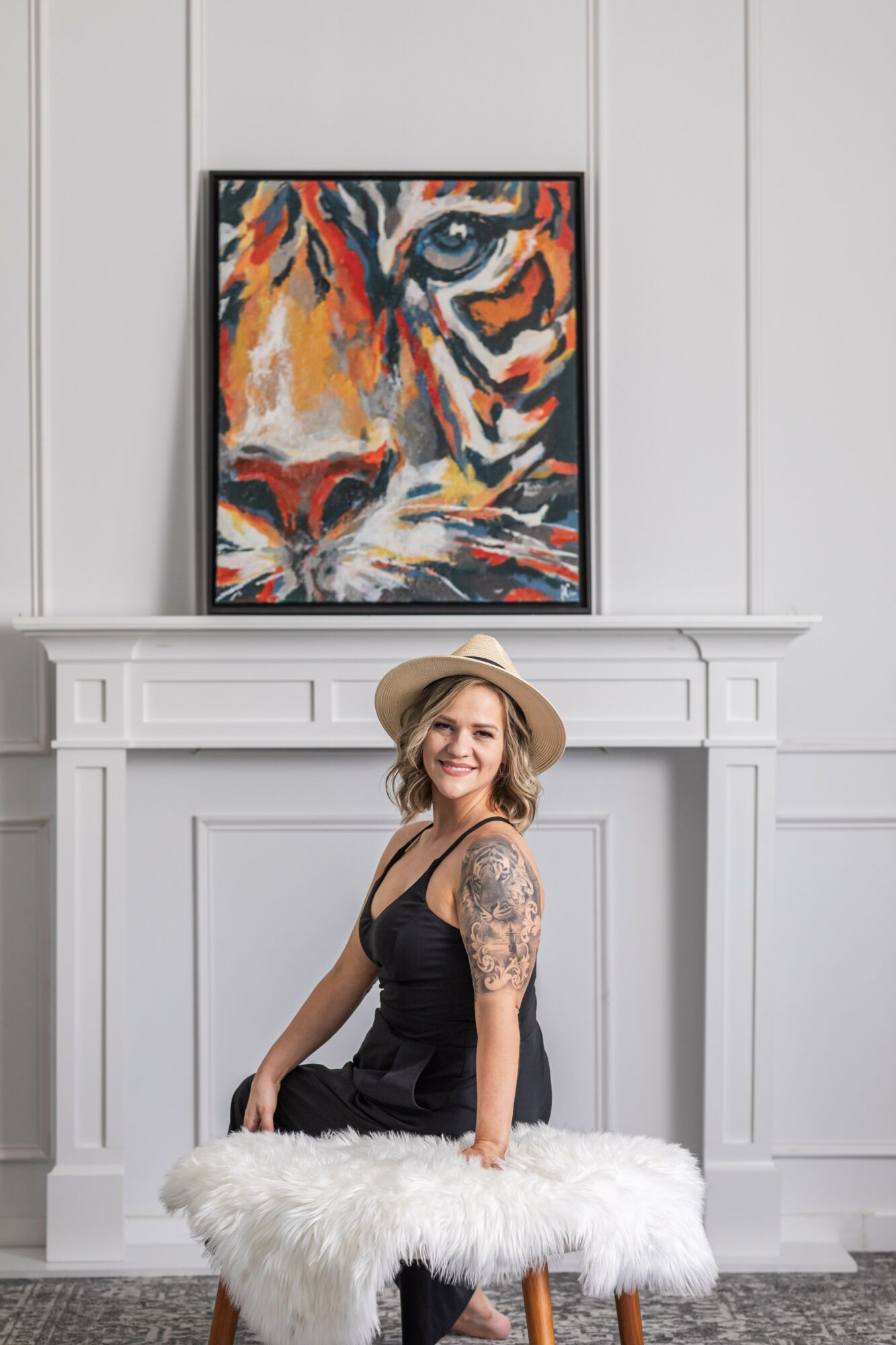 Woman with a hat and tattooed arm sitting on a white furry stool in front of a fireplace, with a tiger painting above.