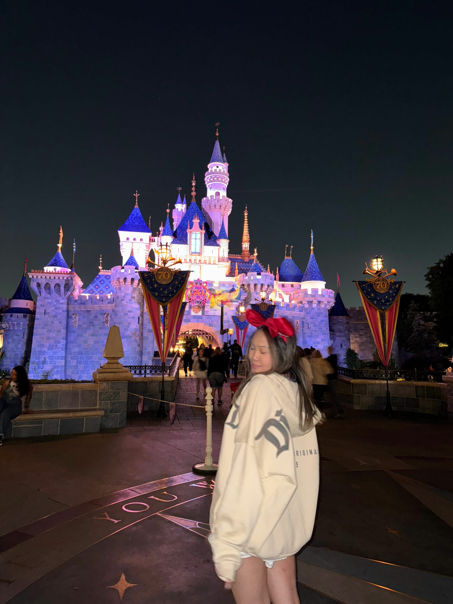 Person in white coat standing in front of a colorful castle at night, illuminated with lights, with a dark sky background.