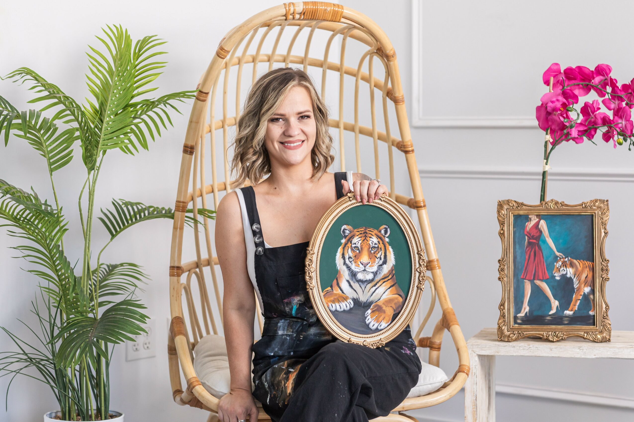 Young woman sitting in a wicker chair holding a framed tiger portrait, surrounded by plants and artwork, smiling.