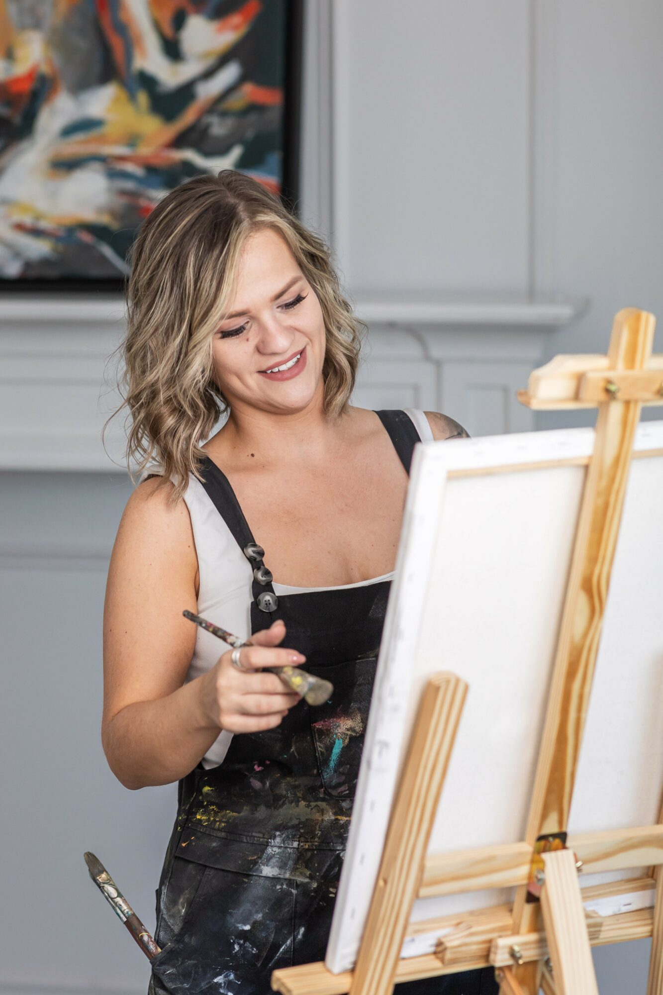 Woman smiling while painting on a canvas in an art studio, holding a palette and brush, with an abstract painting in the background.
