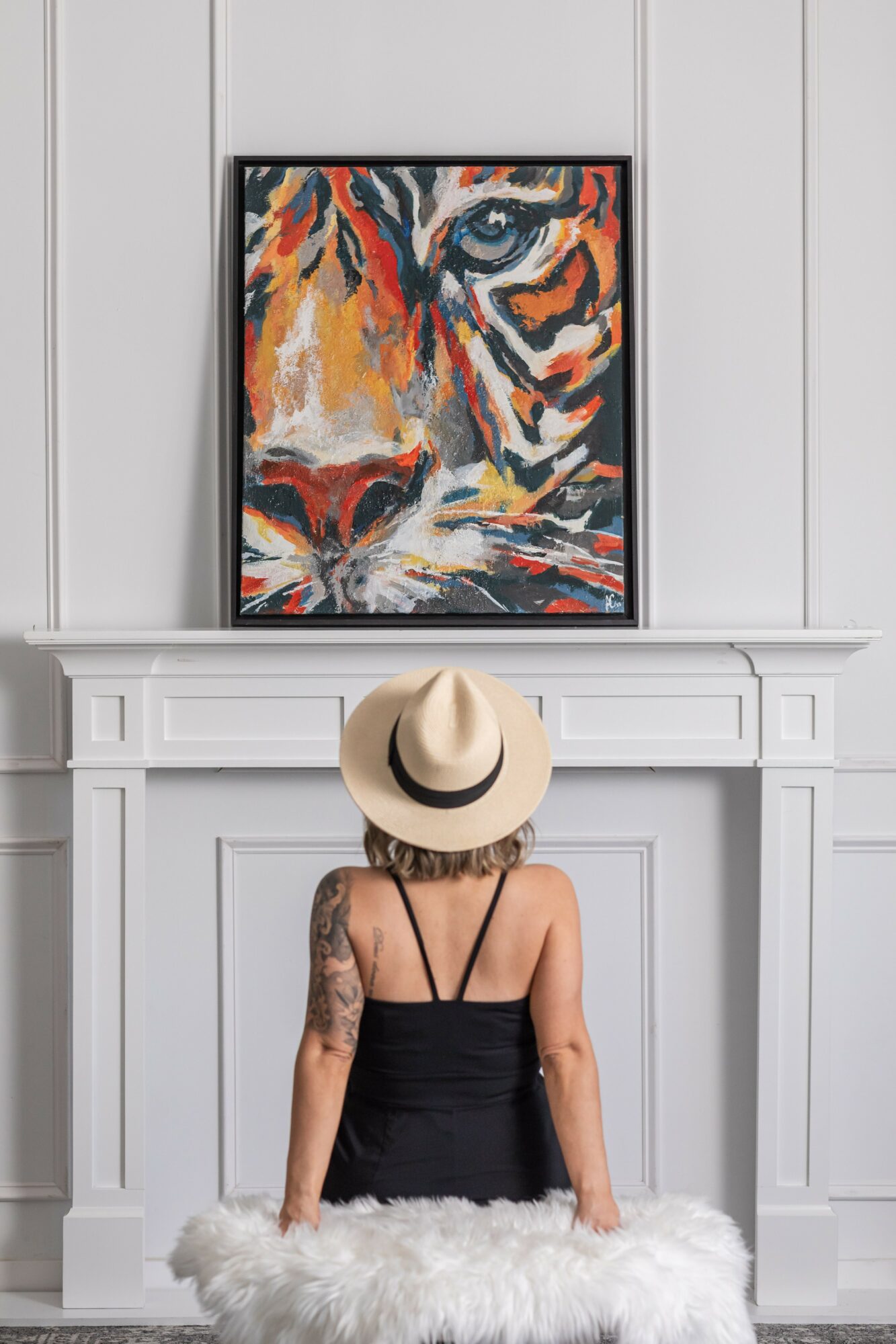Woman with a hat and tattooed arm sitting on a white furry cushion in front of a white fireplace, looking at a tiger painting.